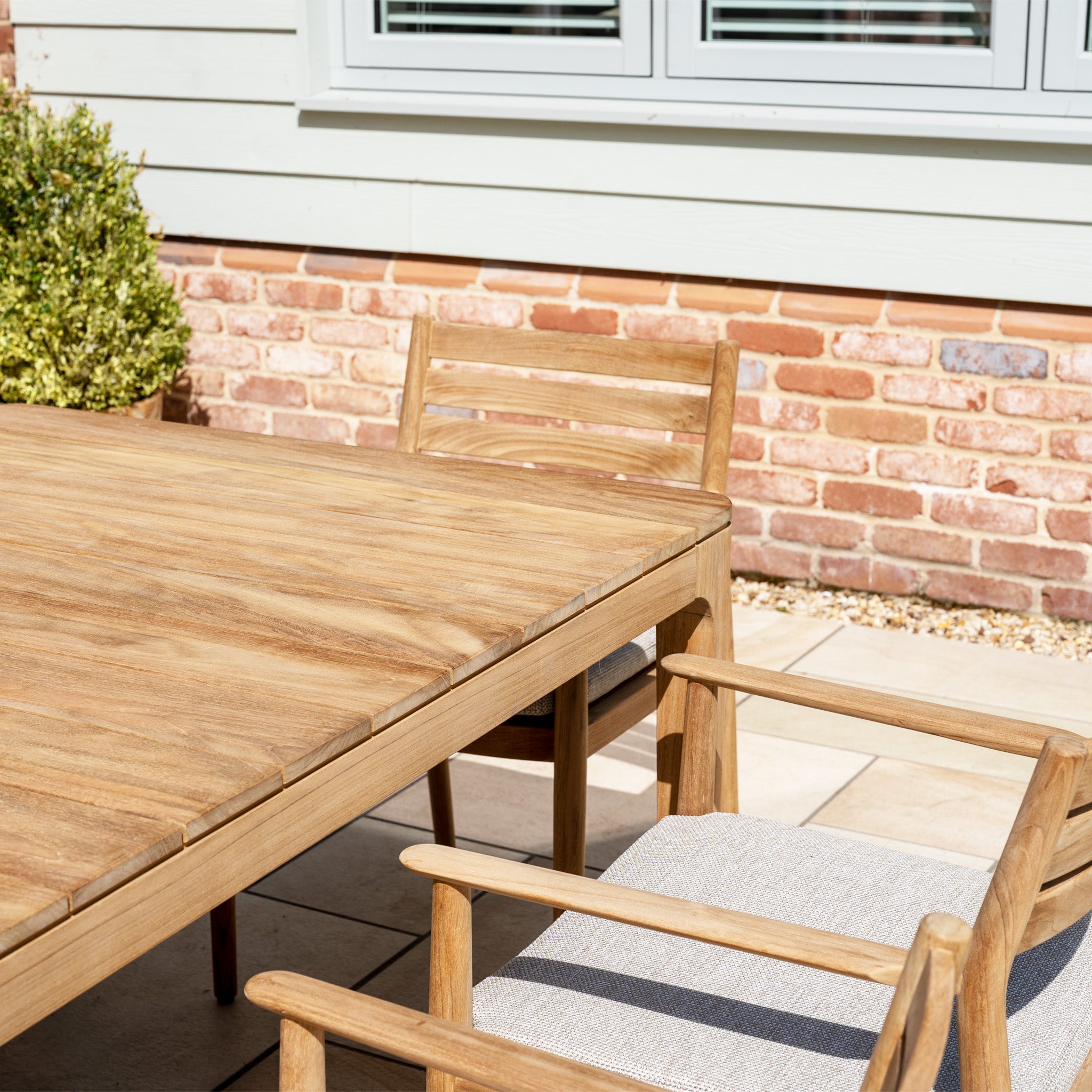 The Siena 8 Seat Dining Set with Teak Table in Fawn is arranged on a patio by a brick wall and window, making for an inviting outdoor dining space.
