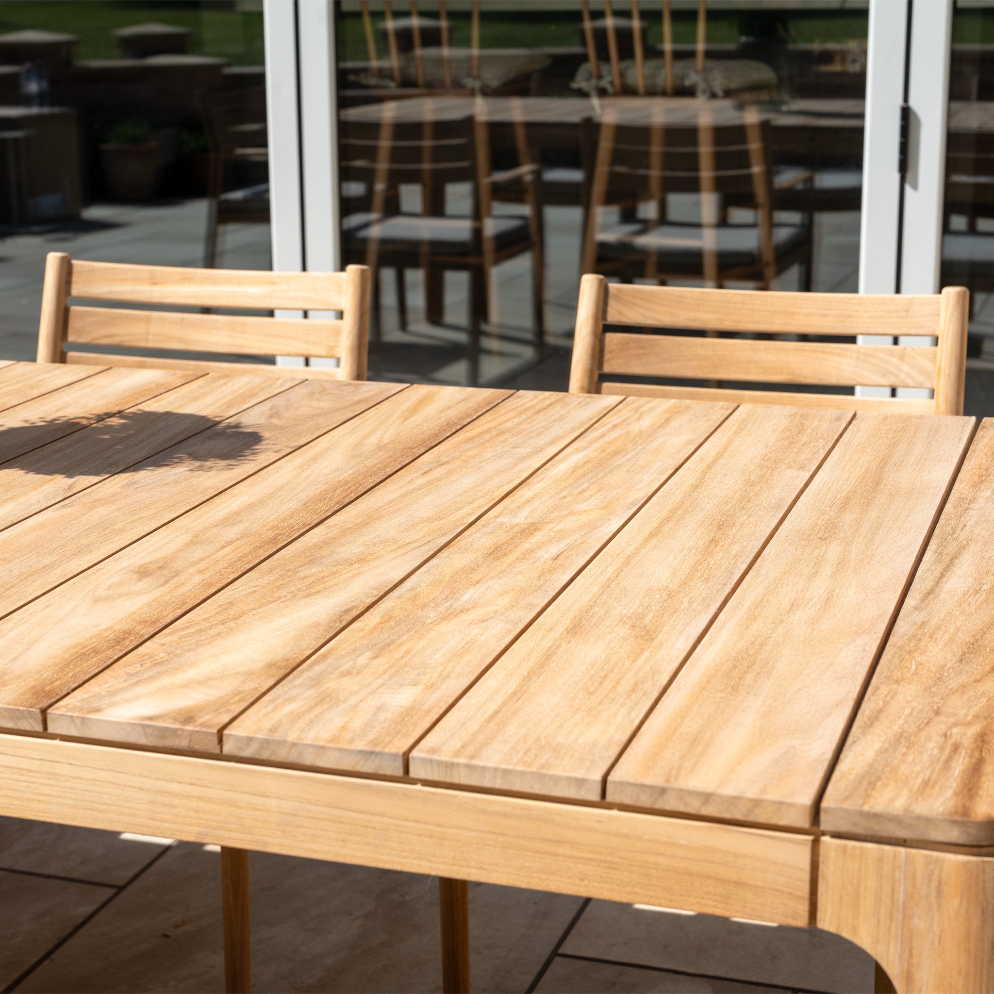 The Siena 8 Seat Dining Set with Teak Table in Fawn enhances a tiled outdoor space, complemented by shimmering reflections in the glass doors behind.