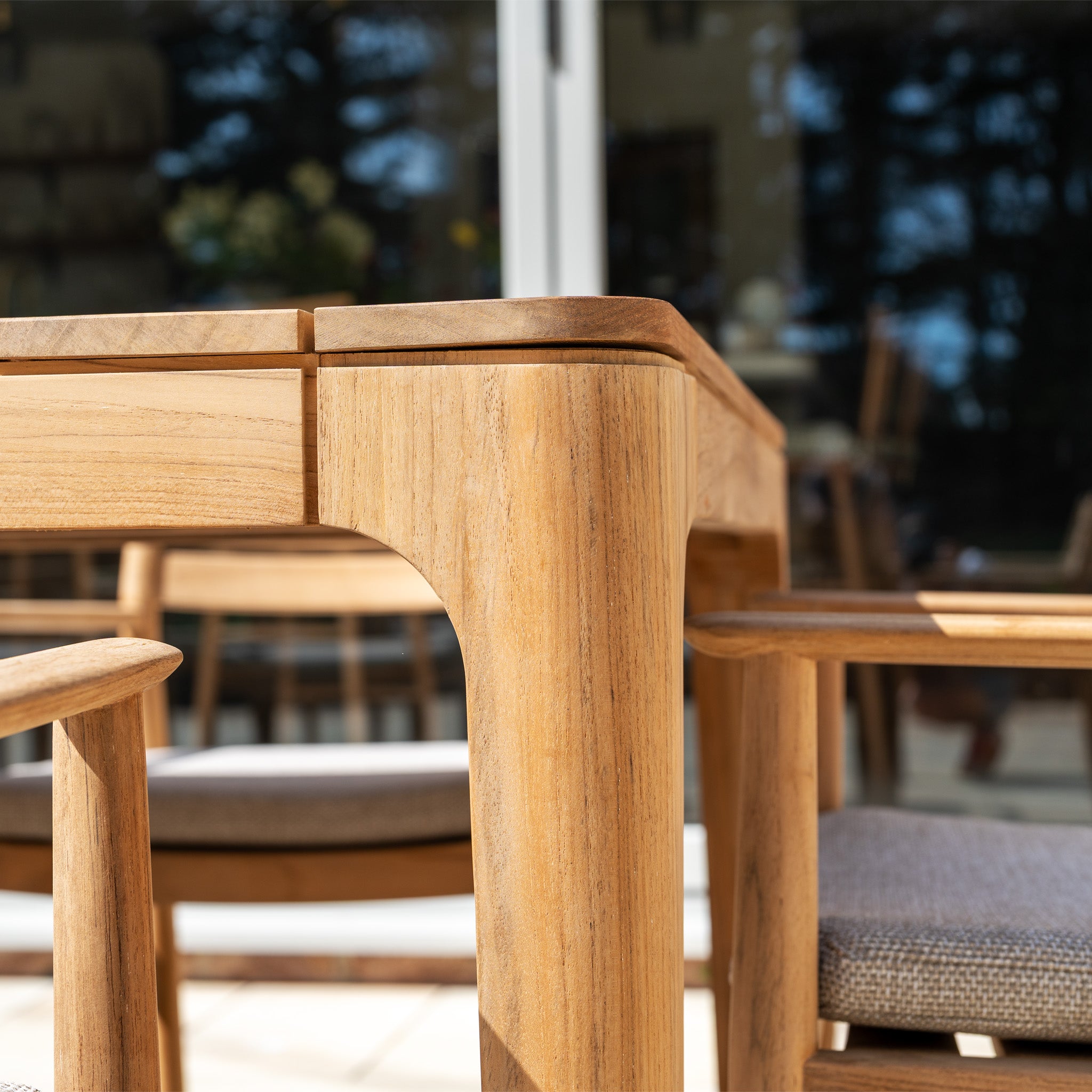 Close-up of the Siena 8 Seat Dining Set with Teak Table in Fawn—wooden table and cushioned chairs arranged outdoors, reflections shimmering in the glass behind.