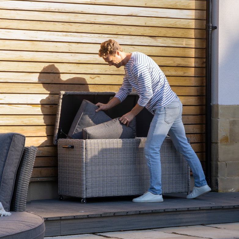 A person in jeans and a striped shirt is organizing cushions into the Pacific Lifestyle - Small Rattan Storage Box in Slate Grey, set on a deck with wood paneling, an essential for garden storage.