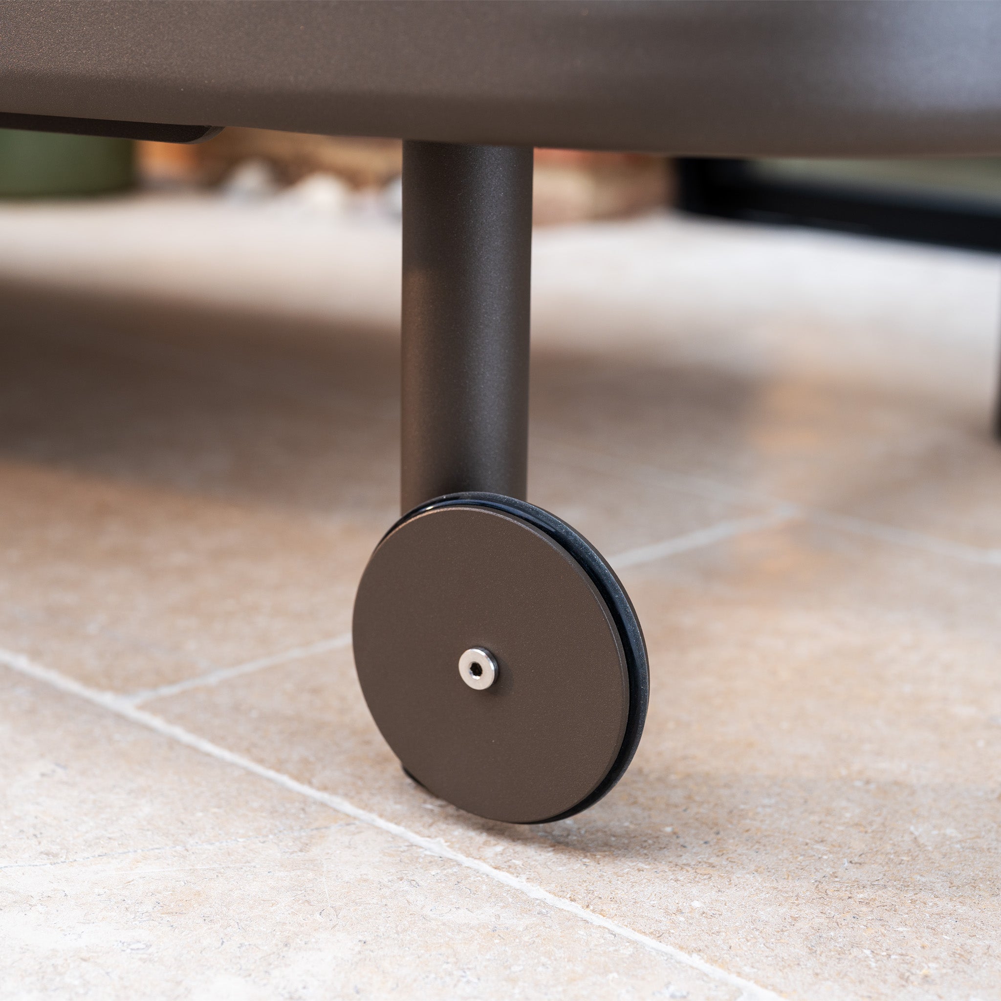 A close-up of a round black wheel on the leg of the Sofia Sunlounger with Headrest and Wheels in Putty, shown on a tiled floor.