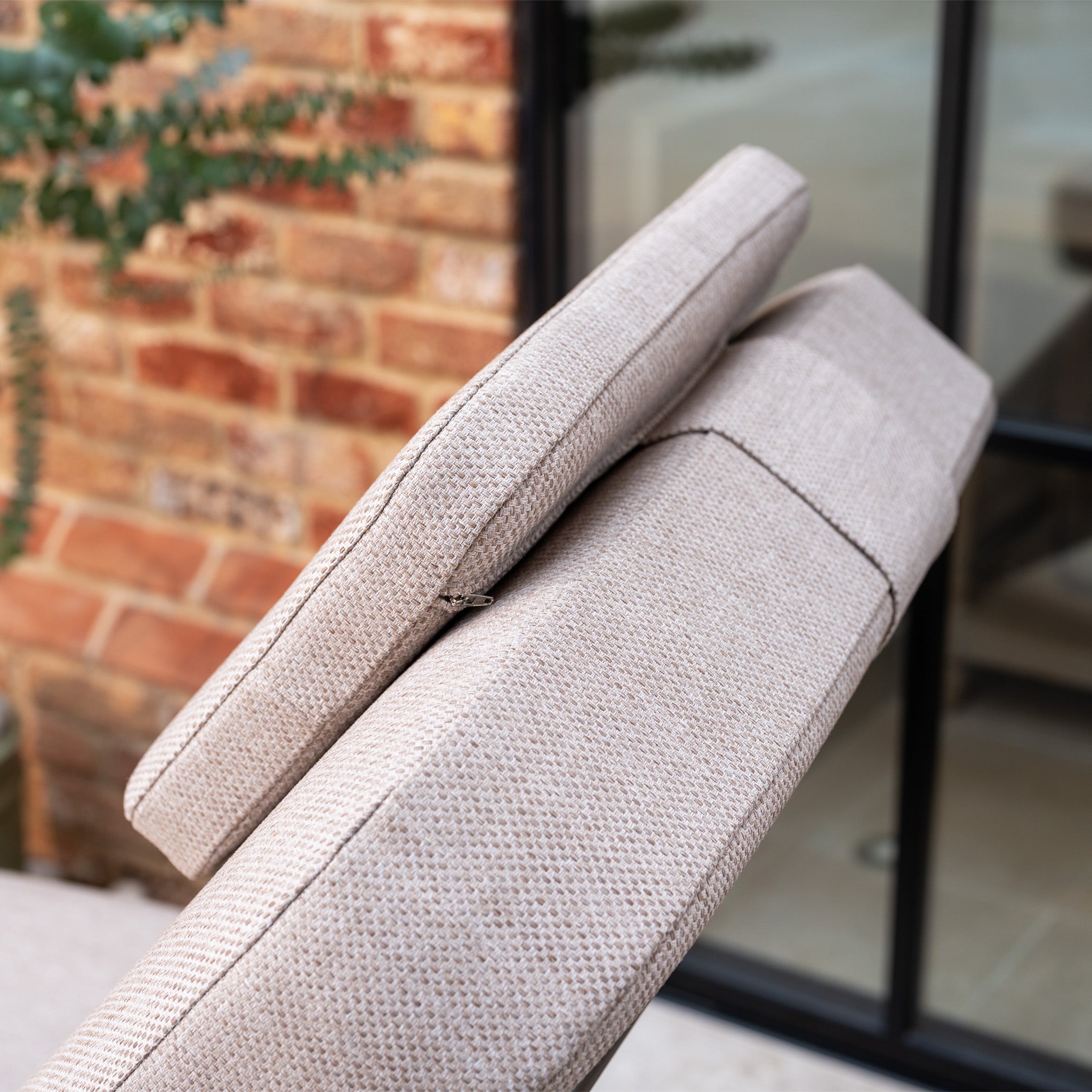 Close-up of adjustable beige fabric sofa cushions by a brick wall and glass door, paired with the Sofia Sunlounger with Headrest and Wheels in Putty—an elegant aluminium sun lounger designed for stylish relaxation.