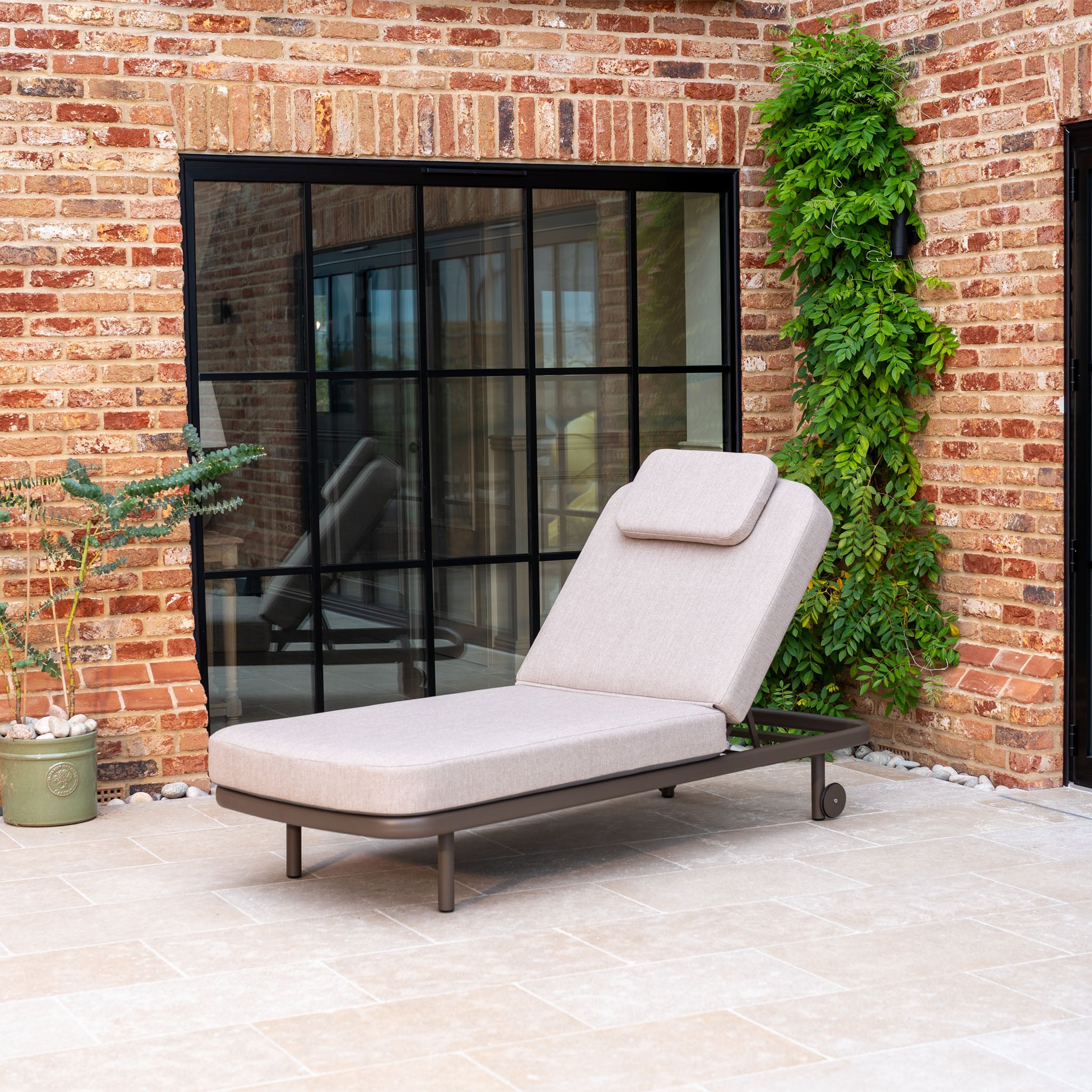 Unwind on the Sofia Sunlounger with Headrest and Wheels in Putty, a chic outdoor lounger perfect for your patio, featuring a comfortable cushion and stylish design amidst garden scenery.