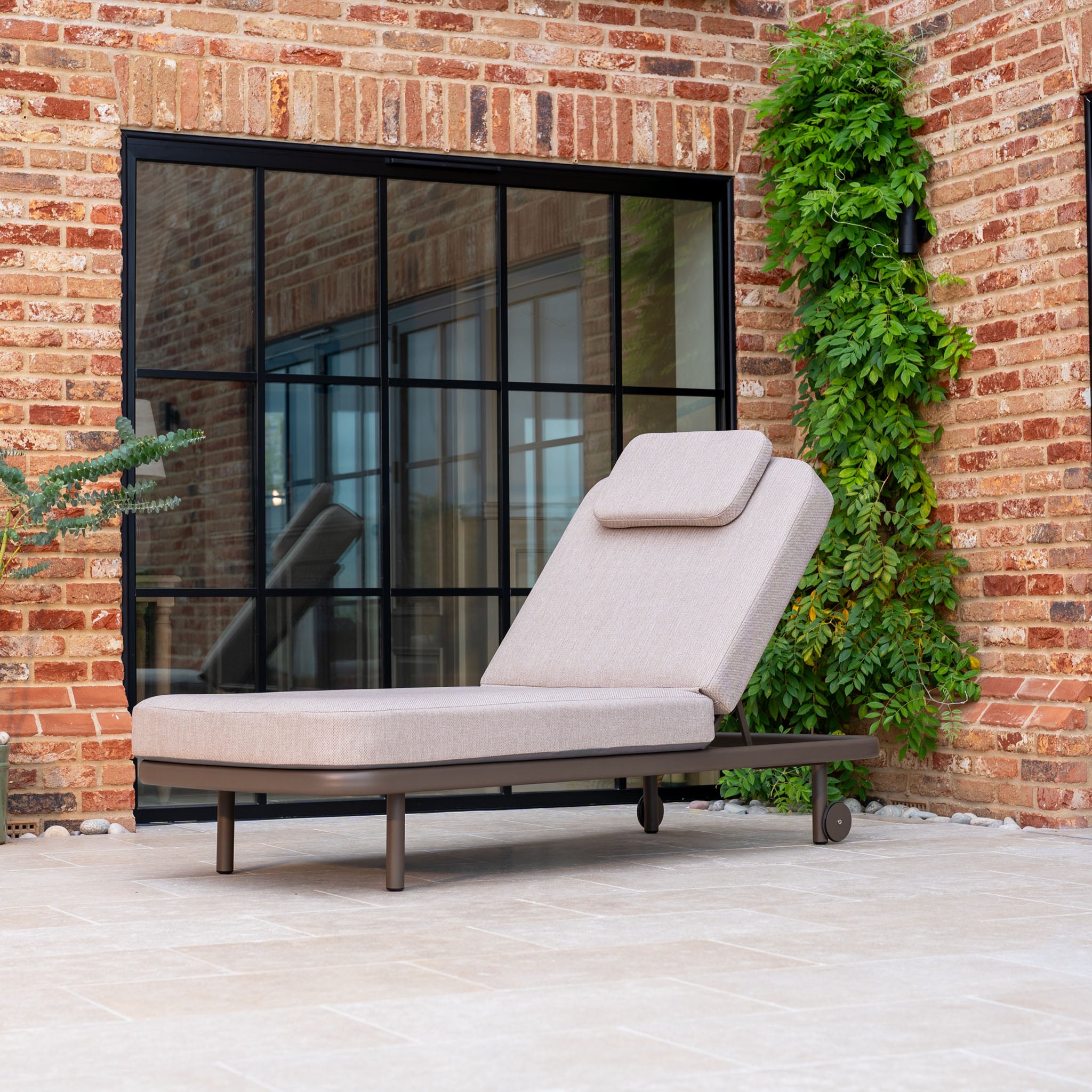 Sofia Sunlounger with Headrest and Wheels in Putty on a patio by a brick wall with a large window and green climbing plant.