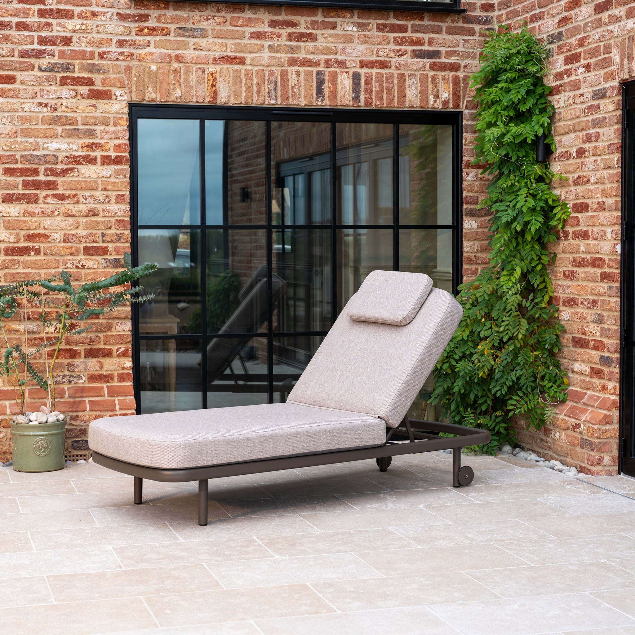 The Sofia Sunlounger with Headrest and Wheels in Putty sits on a tiled patio by a brick wall and window, surrounded by greenery.