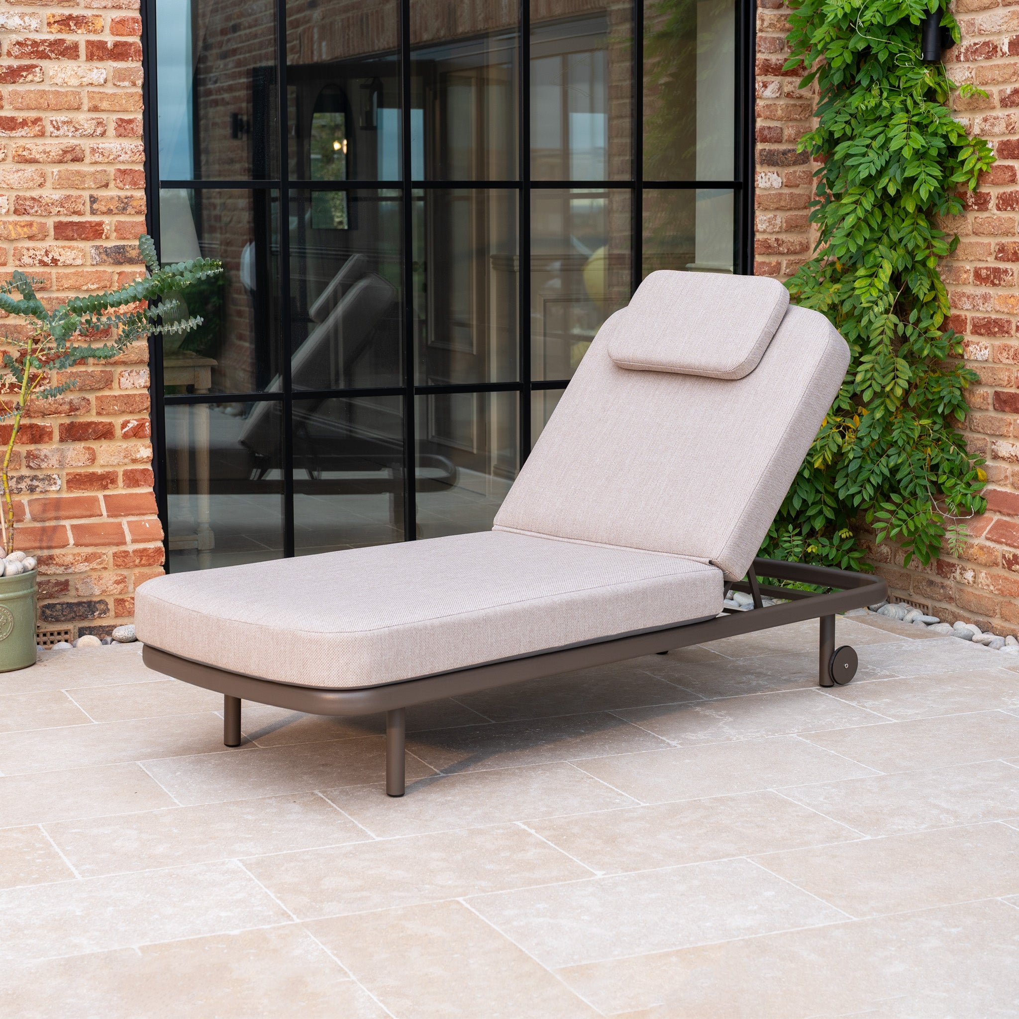 The Sofia Sunlounger with Headrest and Wheels in Putty is an outdoor lounge chair featuring an adjustable backrest, shown on a tiled patio beside a glass wall and a brick wall.