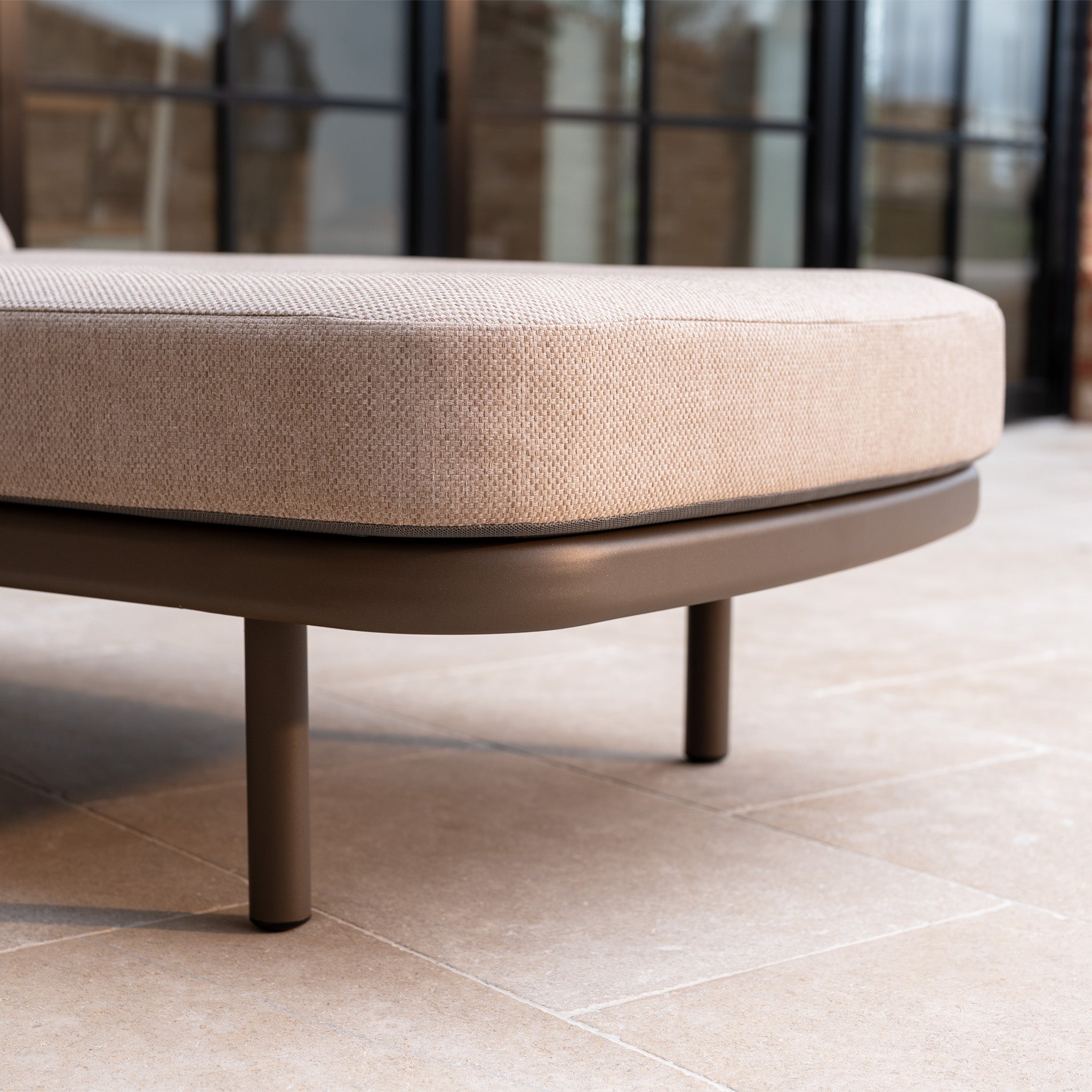 Close-up of a Sofia Sunlounger with Headrest and Wheels in Putty on a tiled patio near glass doors, featuring brown legs.