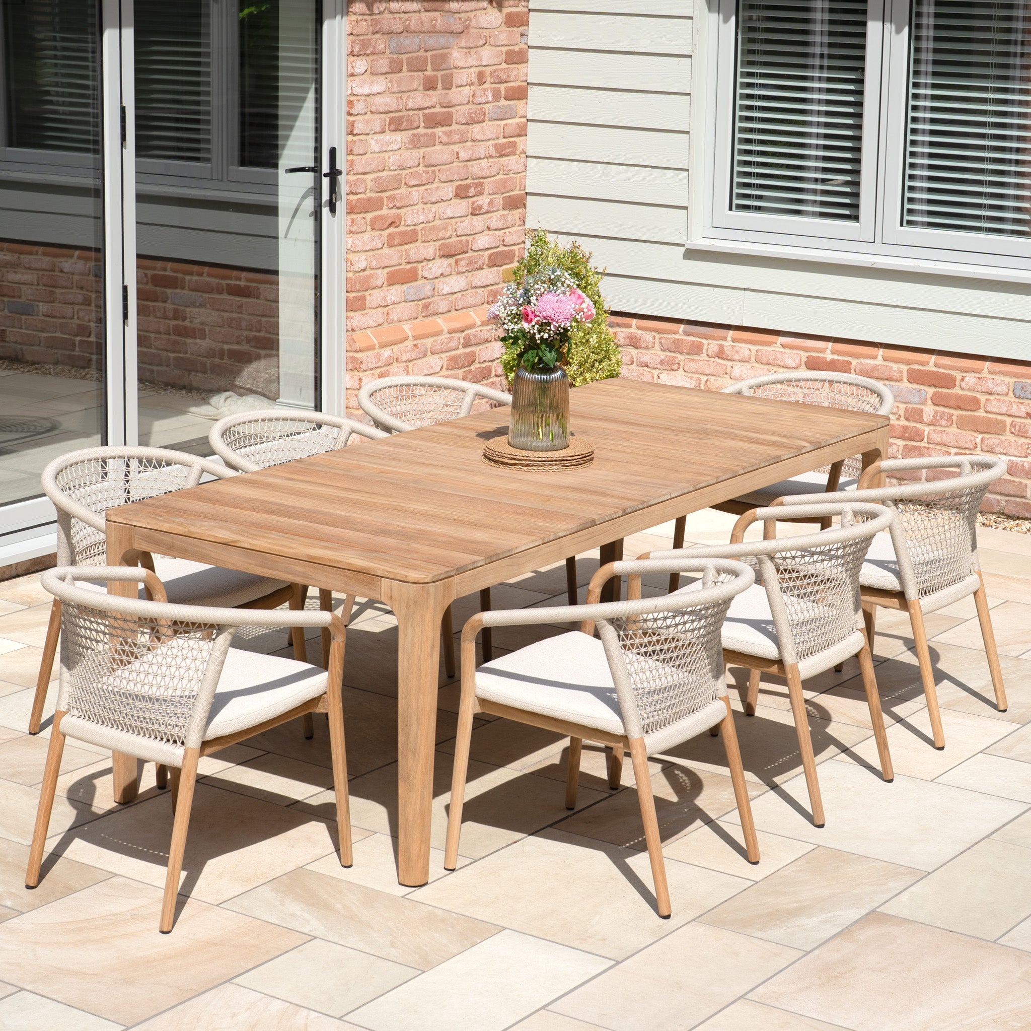 The Sora 8 Seat Rope Dining Set with Teak Table in Stone features eight chairs with handwoven rope details and a wooden table, styled with a flower vase on a tiled patio.