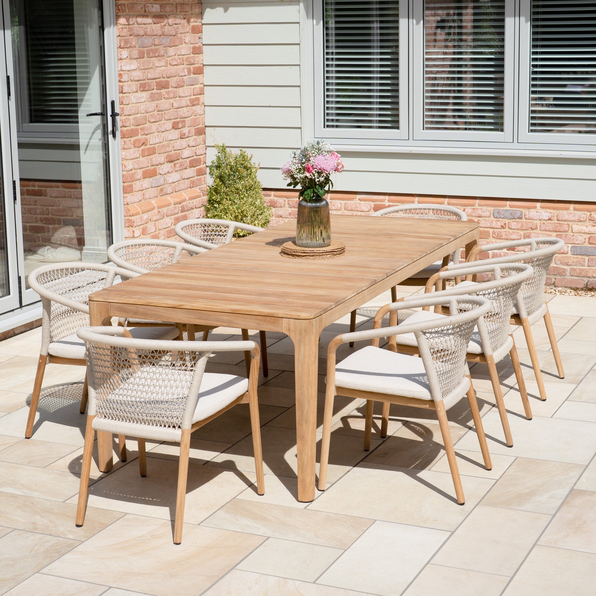 The Sora 8 Seat Rope Dining Set with Teak Table in Stone, complete with eight chairs, forms an inviting outdoor dining area on a tiled patio near the house.