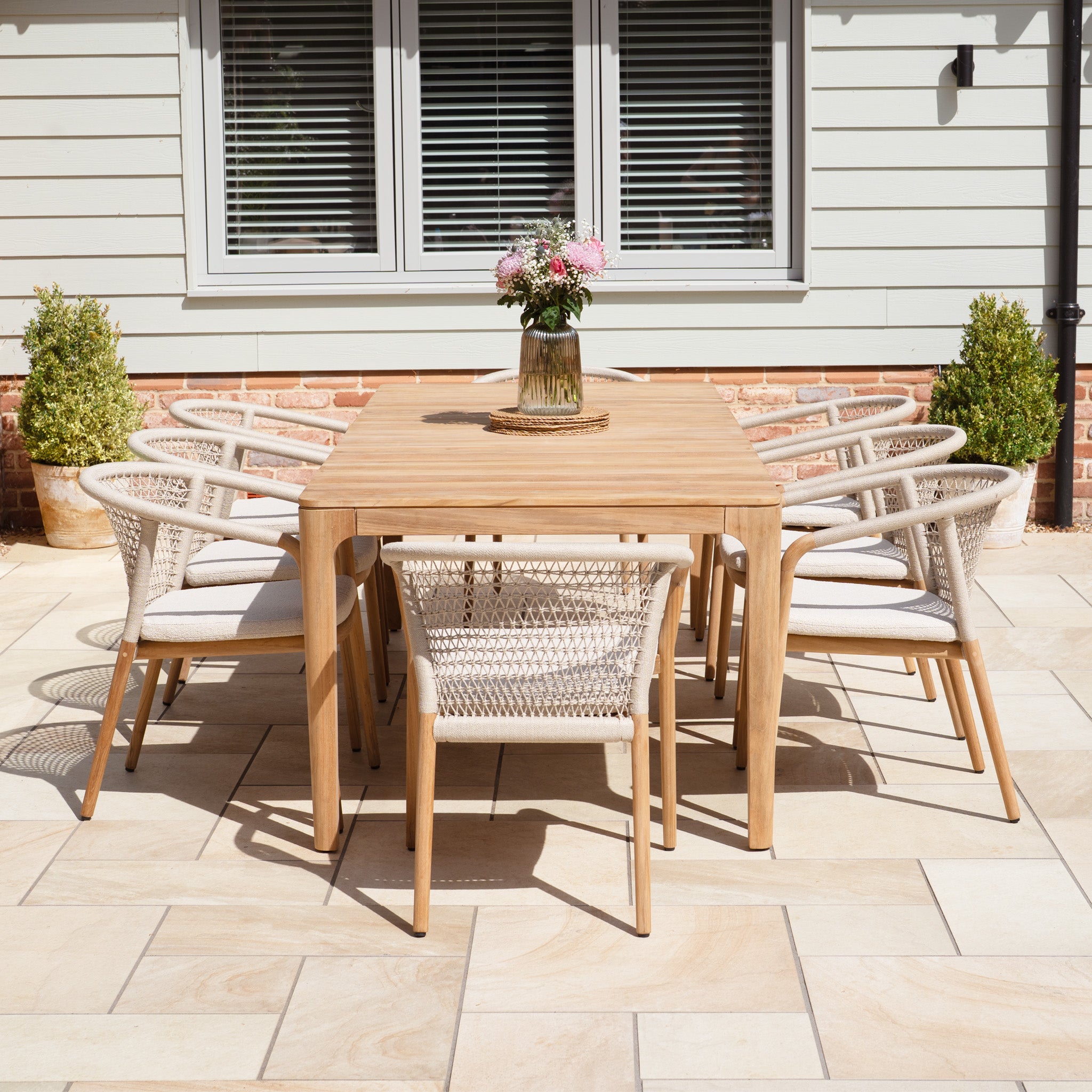 The Sora 8 Seat Rope Dining Set with Teak Table in Stone features a stylish teak table and eight rope-detailed chairs, perfect for outdoor dining on a sunlit patio.