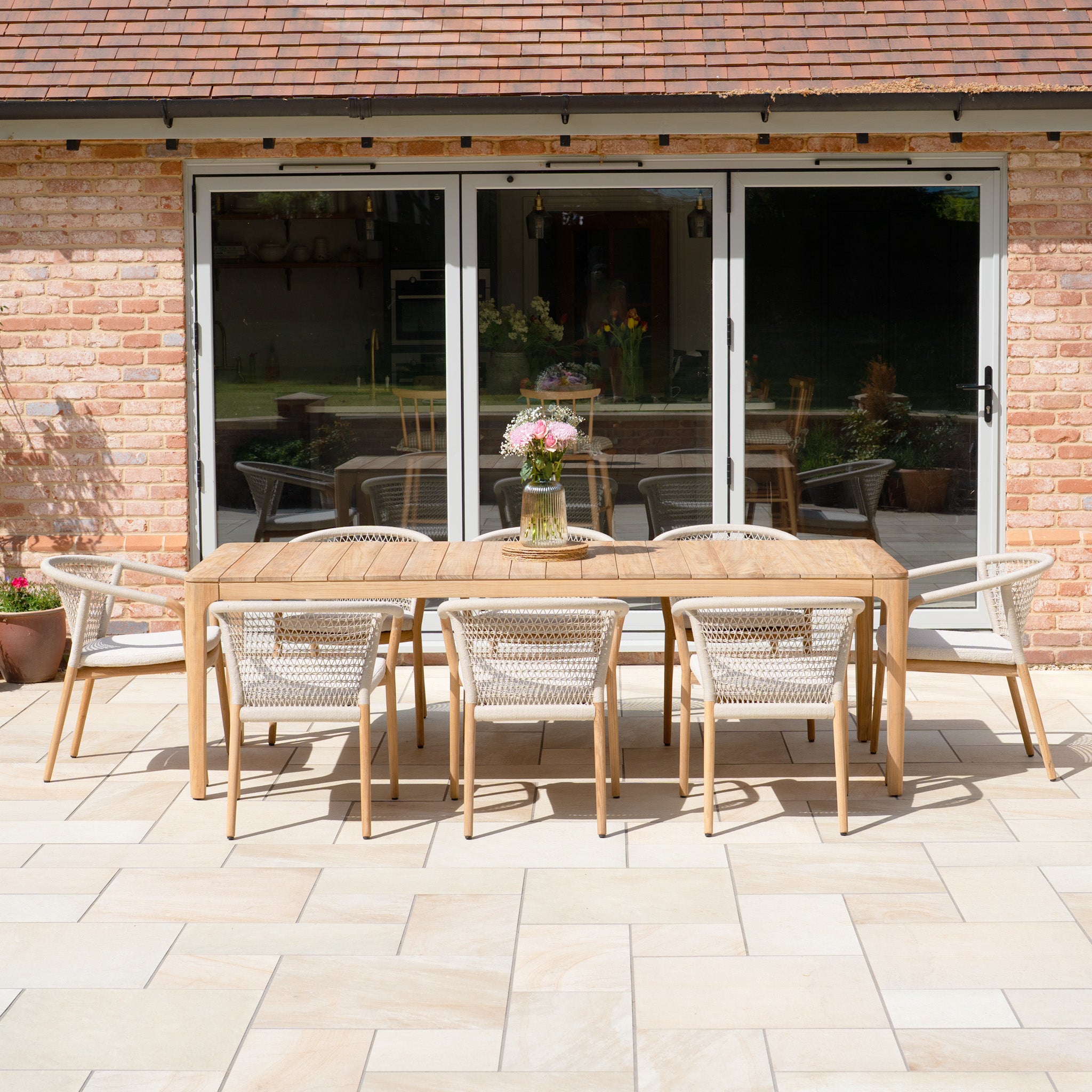 The Sora 8 Seat Rope Dining Set with Teak Table in Stone, featuring handwoven rope and seating for eight, is arranged on a stone patio near glass doors with a flower vase centerpiece.