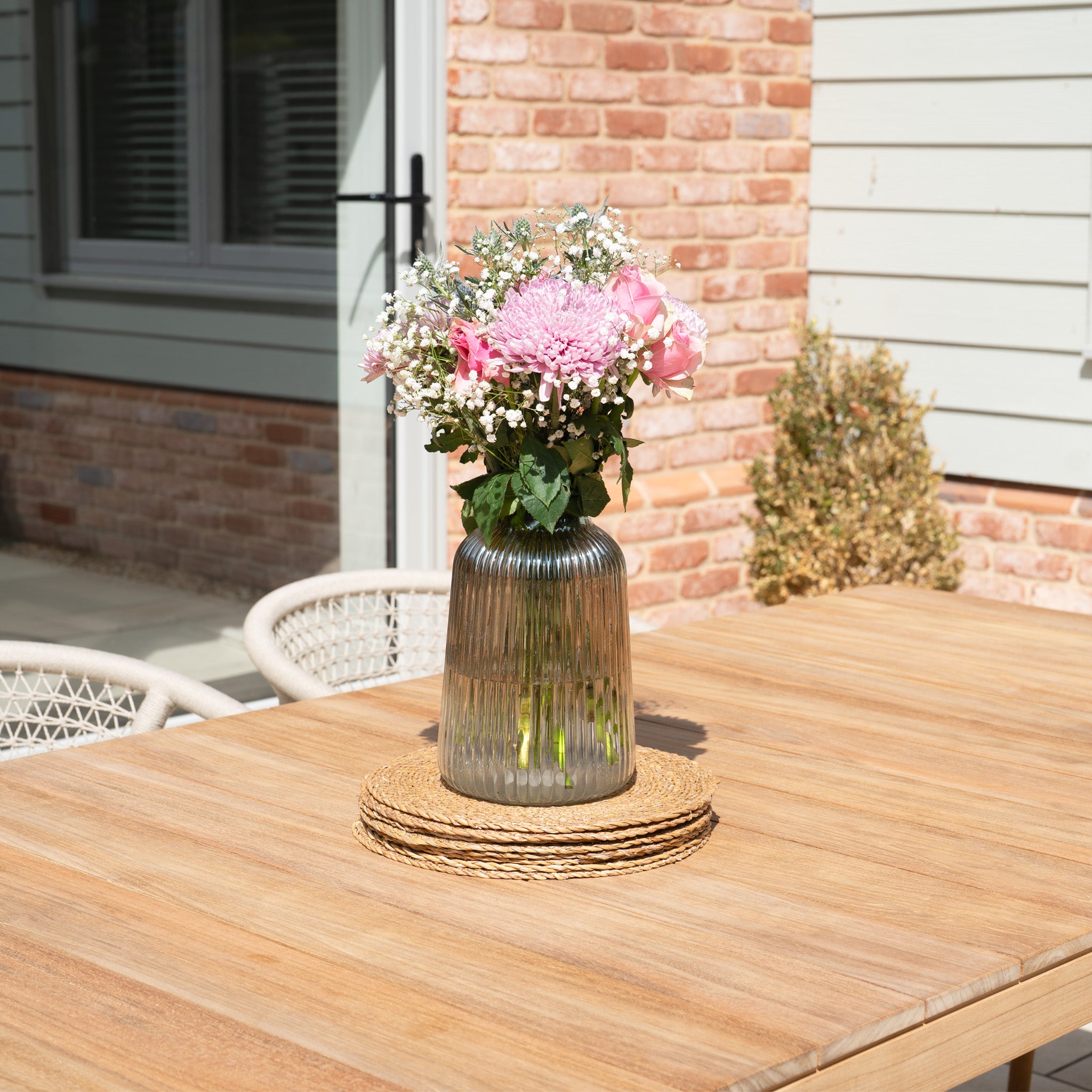 A glass vase with pink flowers sits on the Sora 8 Seat Rope Dining Set with Teak Table in Stone, placed outdoors in the sunlight.