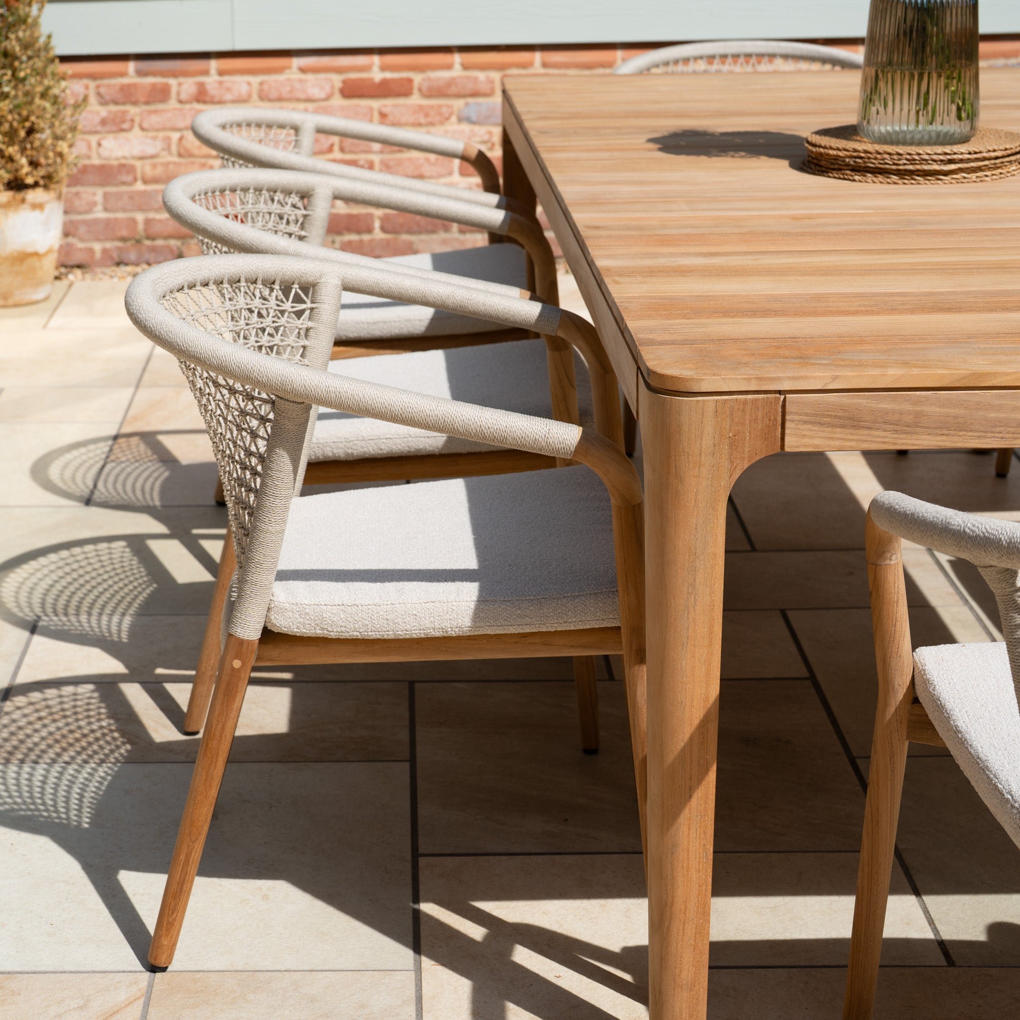 The Sora 8 Seat Rope Dining Set with Teak Table in Stone features weather-resistant materials and light cushioned chairs, creating elegant shadows across your tiled patio.
