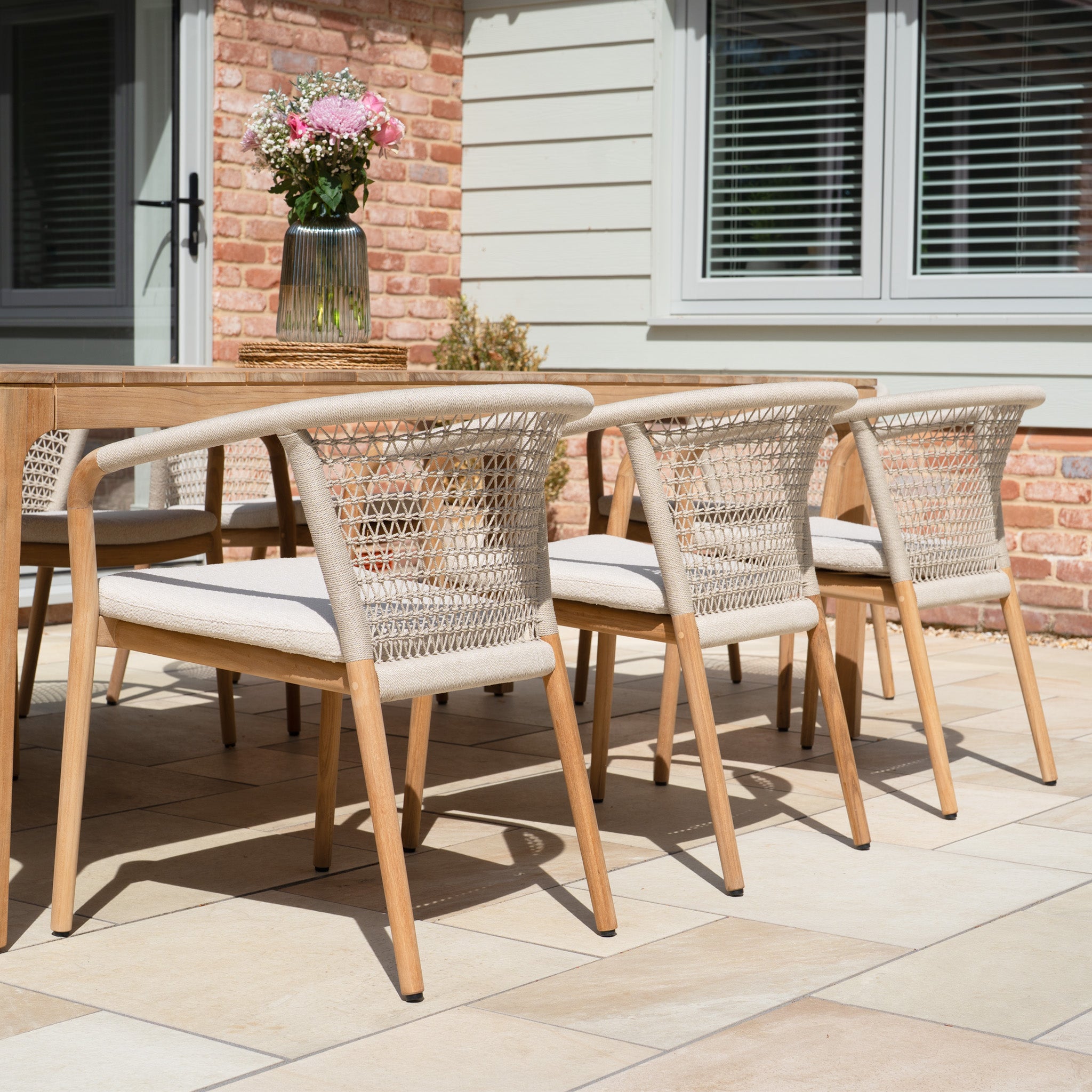 The Sora 8 Seat Rope Dining Set with Teak Table in Stone features weather-resistant rope chairs and a teak table, seating eight for stylish outdoor dining. It's perfect for any patio gathering and topped with a decorative vase of flowers.