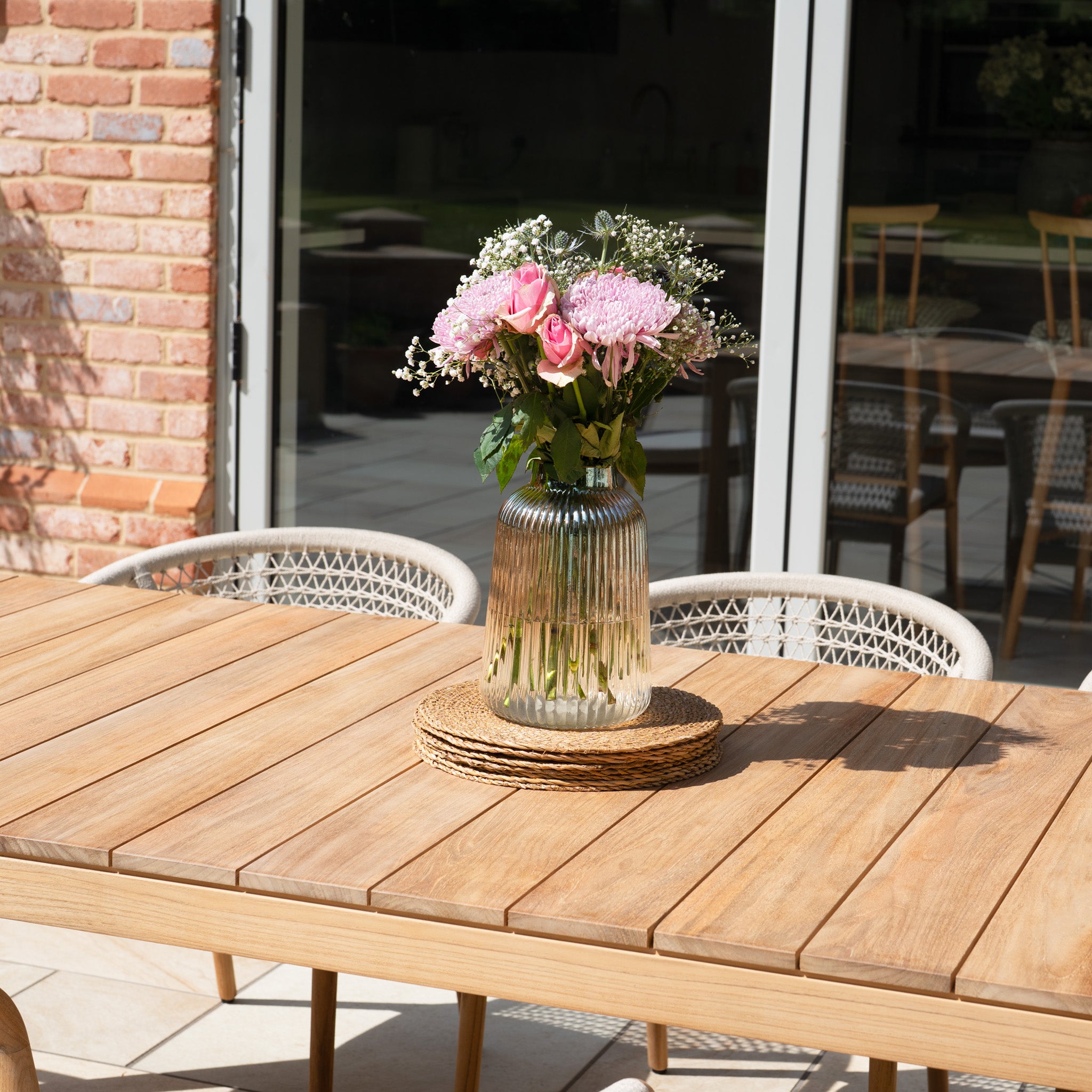 A vase with pink flowers sits on the Sora 8 Seat Rope Dining Set with Teak Table in Stone, basking in sunlight near glass doors.