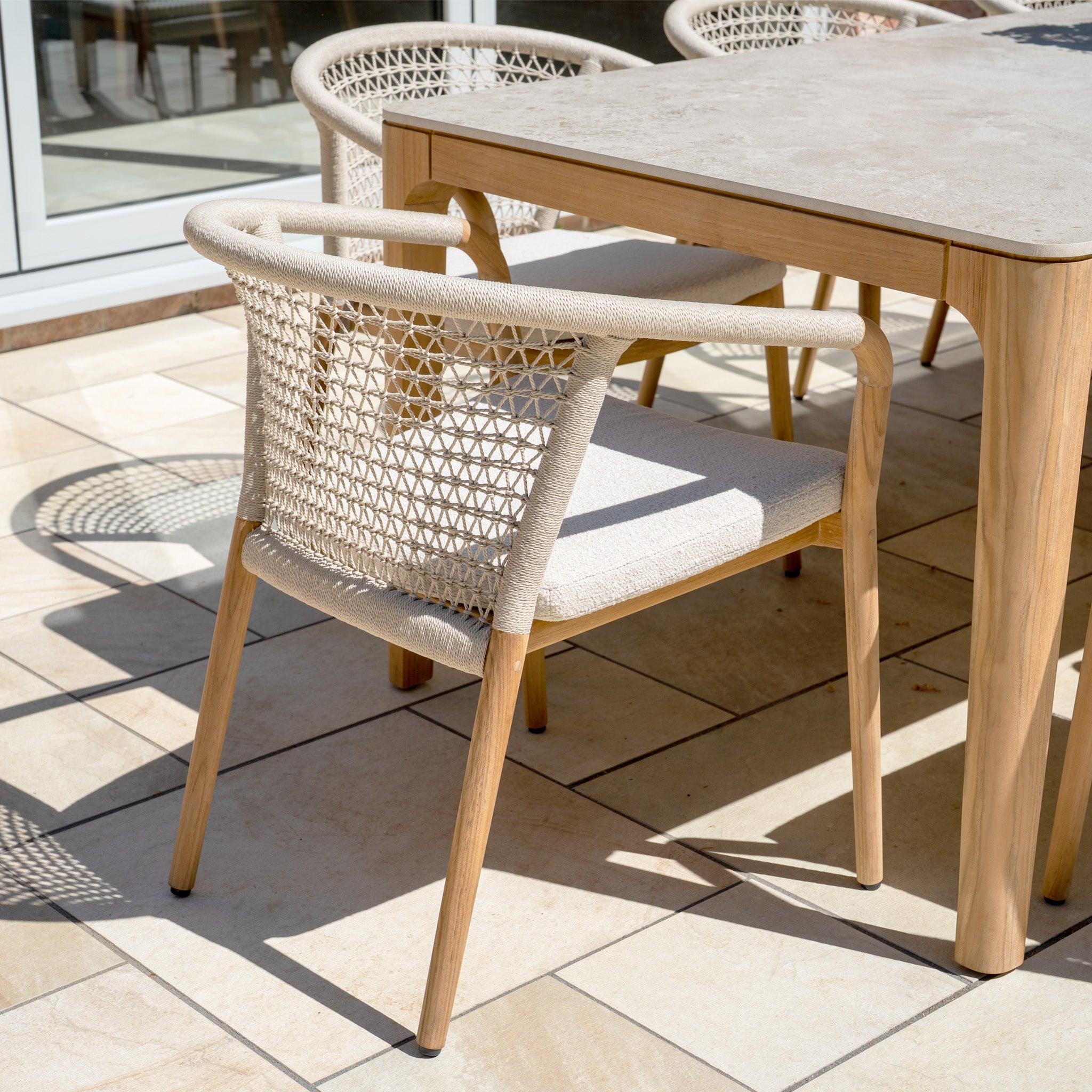 The Sora 8 Seat Rope Dining Set with Ceramic Top Table in Stone sits on a sunlit tiled patio, featuring elegant chairs and a stylish table for outdoor dining.