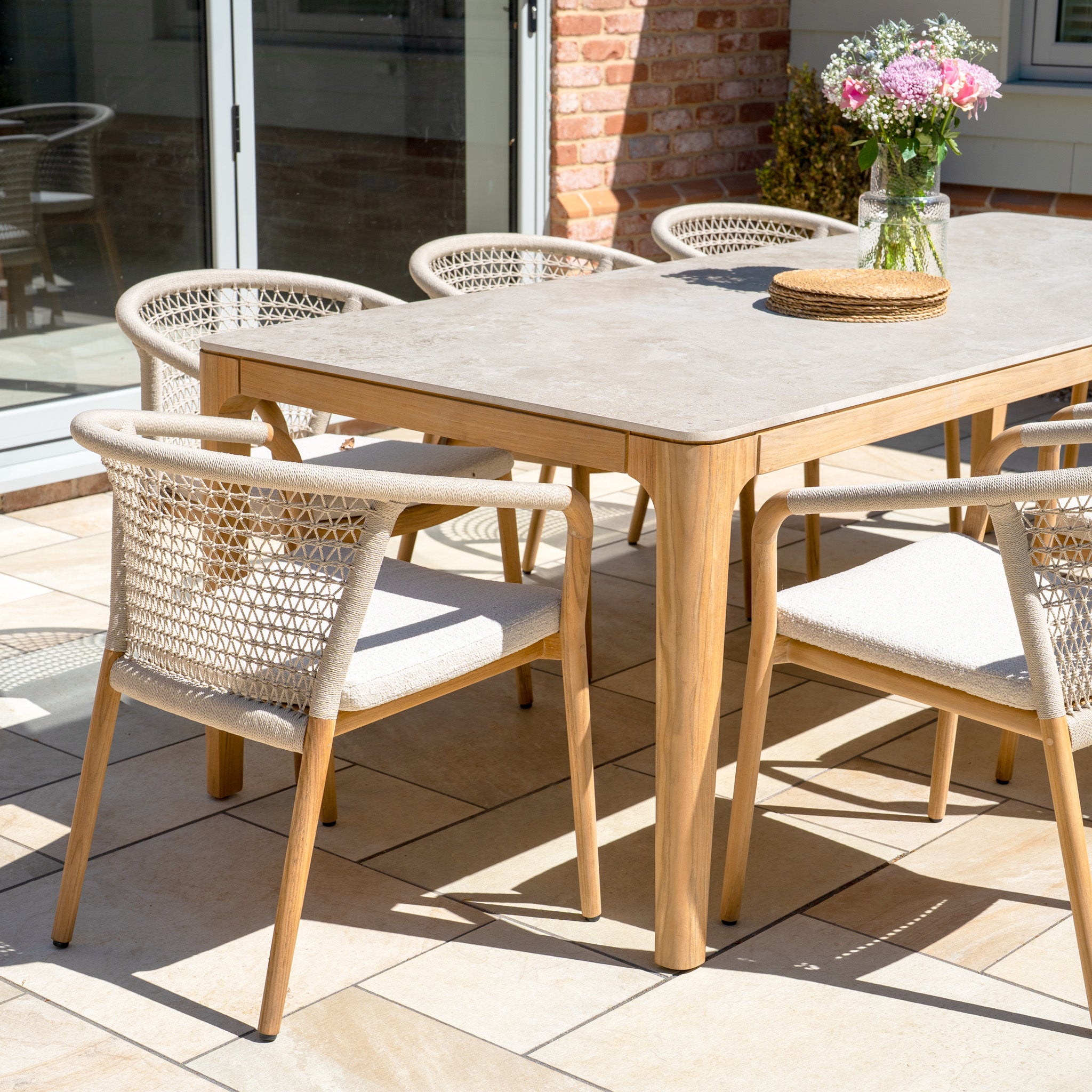 The Sora 8 Seat Rope Dining Set features a ceramic top table in Stone and eight woven chairs, perfect for outdoor dining on a sunlit patio.