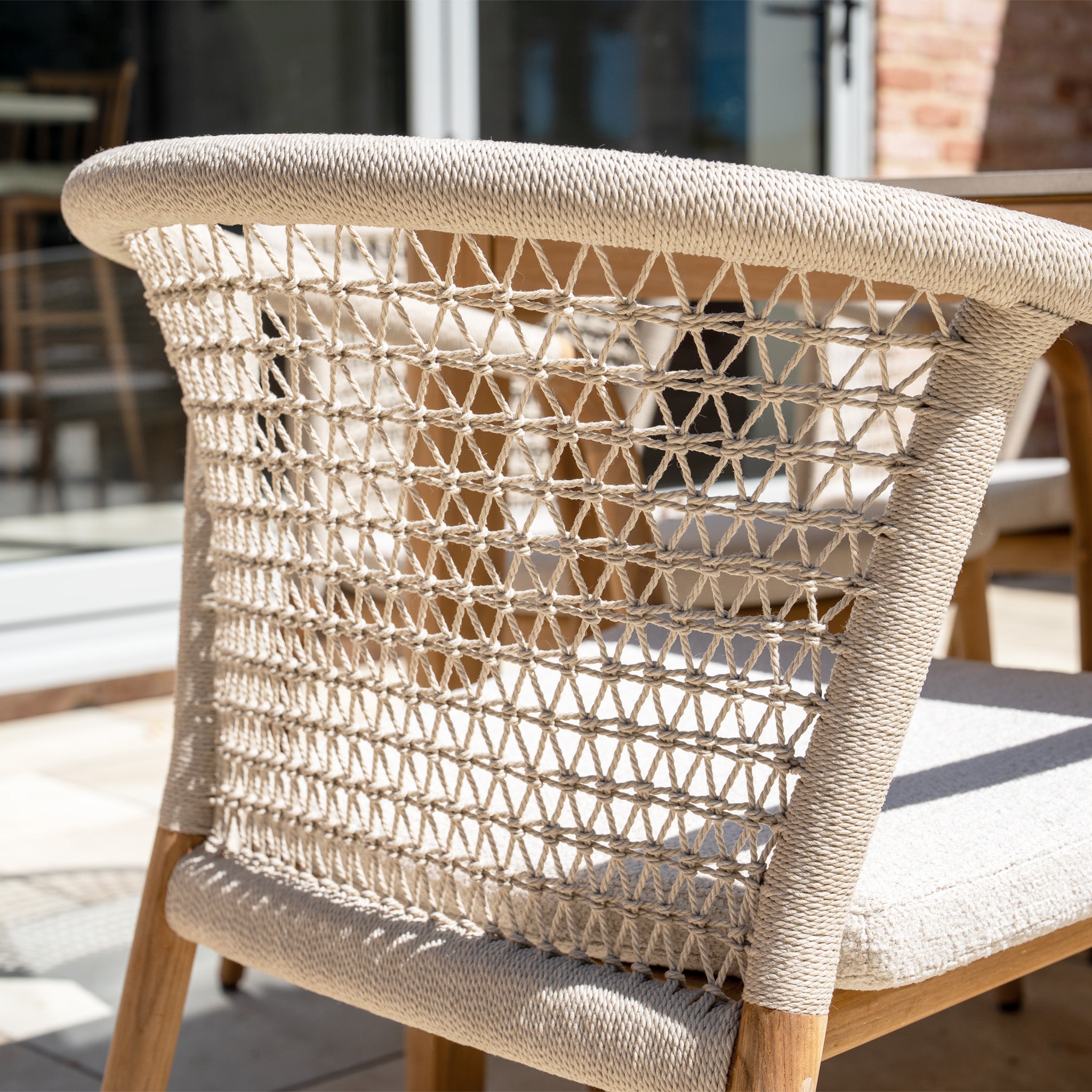 A close-up of the Sora 8 Seat Rope Dining Set with a ceramic top table in Stone, featuring woven backrests and light cushions, arranged on a sunny patio.
