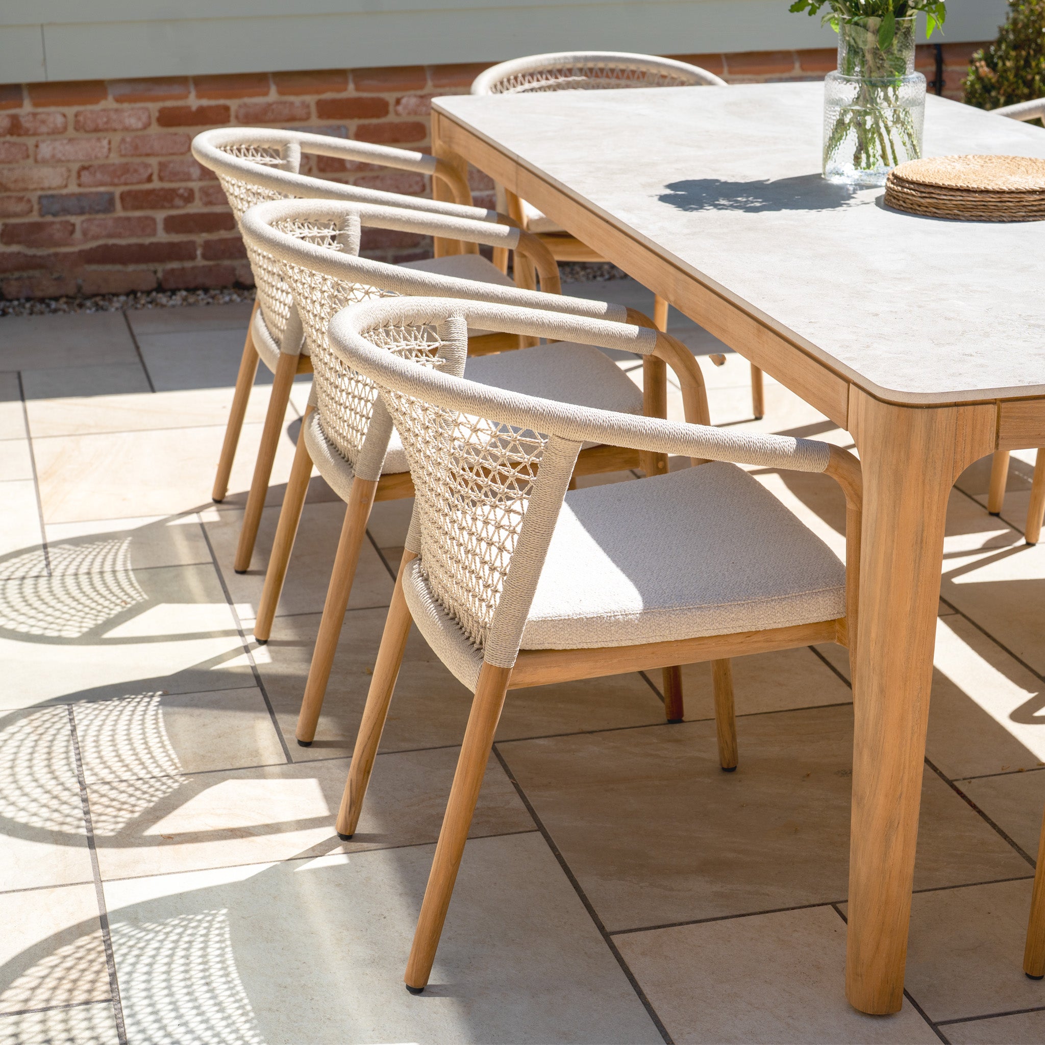 The Sora 8 Seat Rope Dining Set in Stone features handwoven rope chairs and a ceramic top table, perfect for outdoor dining on a tiled patio, accented by a vase of flowers.