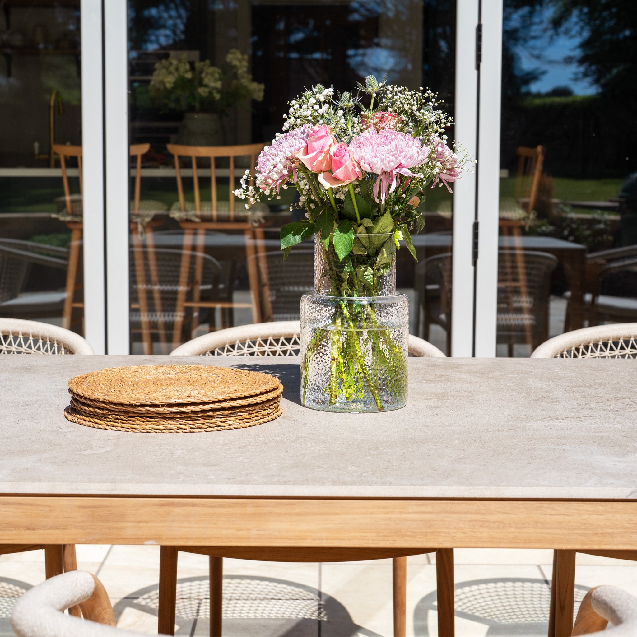 A vase of pink flowers and stacked placemats sit on the Sora 8 Seat Rope Dining Set with Ceramic Top Table in Stone, its handwoven rope detailing and stone finish shining in the sunlight.
