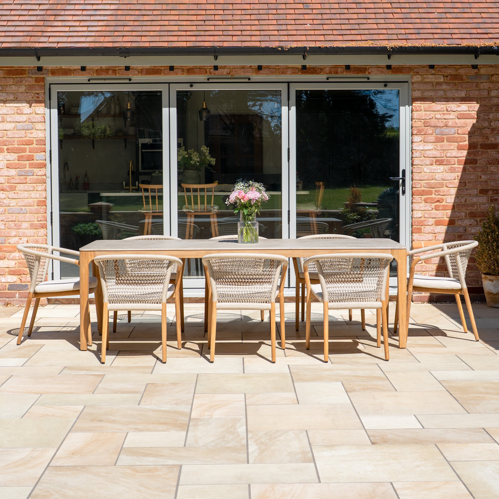The Sora 8 Seat Rope Dining Set with Ceramic Top Table in Stone includes a stylish table and eight chairs with handwoven rope accents, beautifully arranged with a floral vase by glass doors.