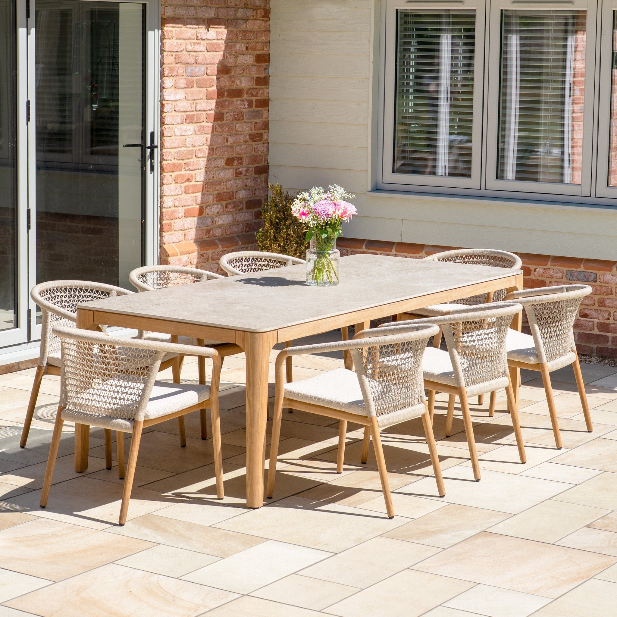 The Sora 8 Seat Rope Dining Set features a ceramic top table in stone and rope-accented chairs, making it perfect for stylish outdoor patio gatherings.