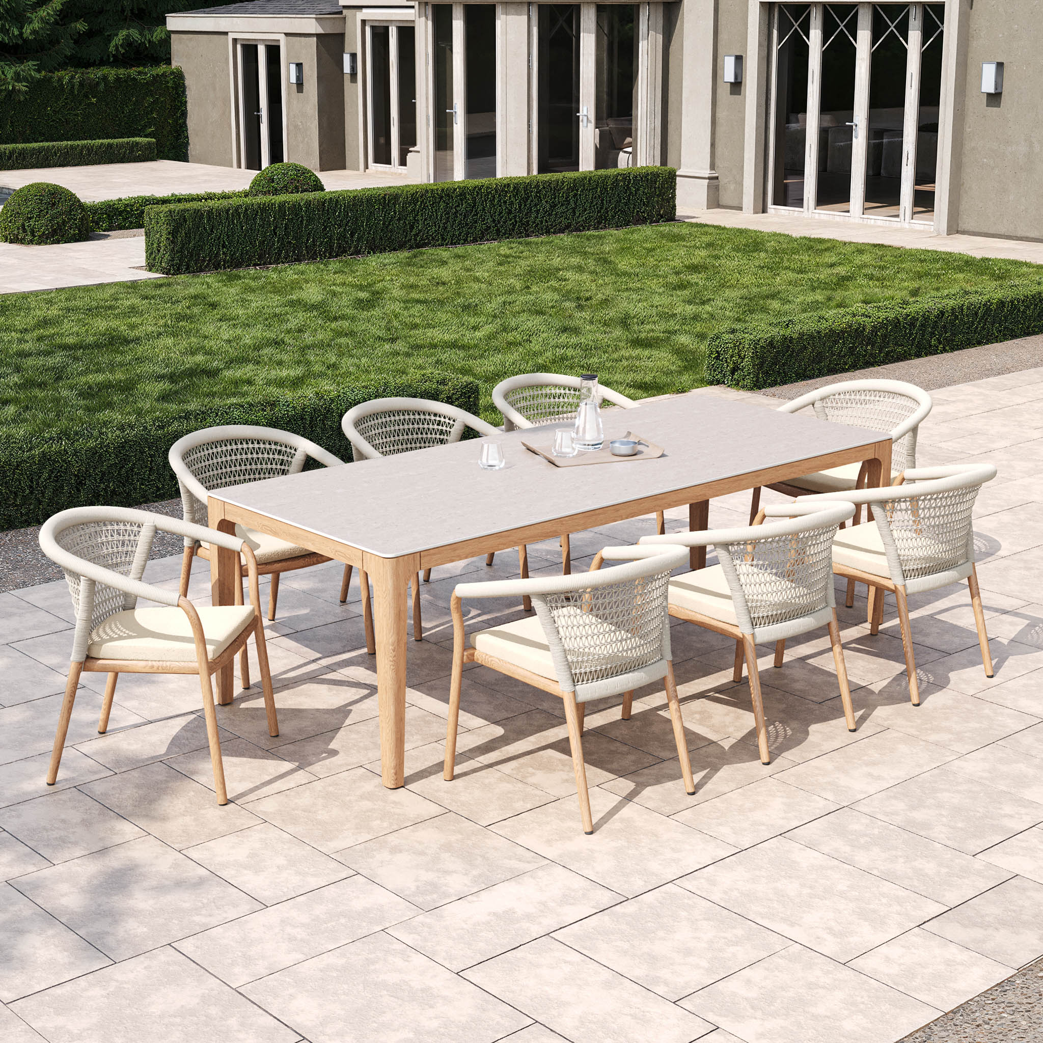 An outdoor dining space highlights the sophisticated Sora 8 Seat Rope Dining Set with Ceramic Top Table in Stone, including a teak frame and woven-back chairs, set on a patio near a green lawn by a modern building.