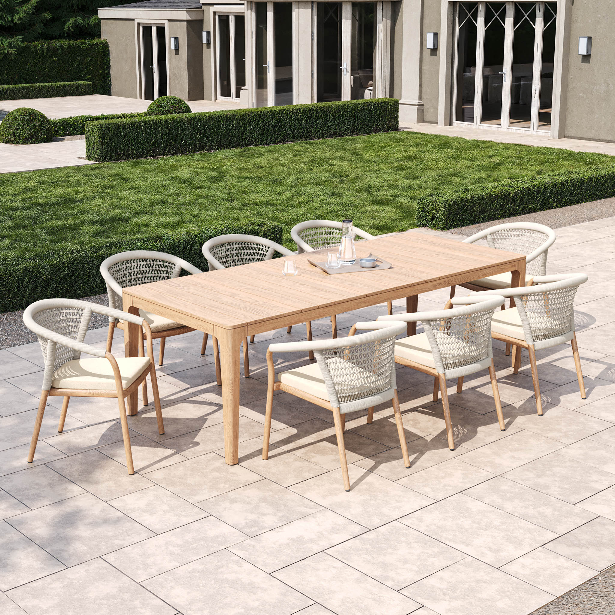 The Sora 8 Seat Rope Dining Set with Teak Table in Stone, set on a tiled patio amid green hedges and grass, is crafted from weather-resistant materials for durability. Its elegant handcrafted rope weave enhances your alfresco dining experience with sophistication.