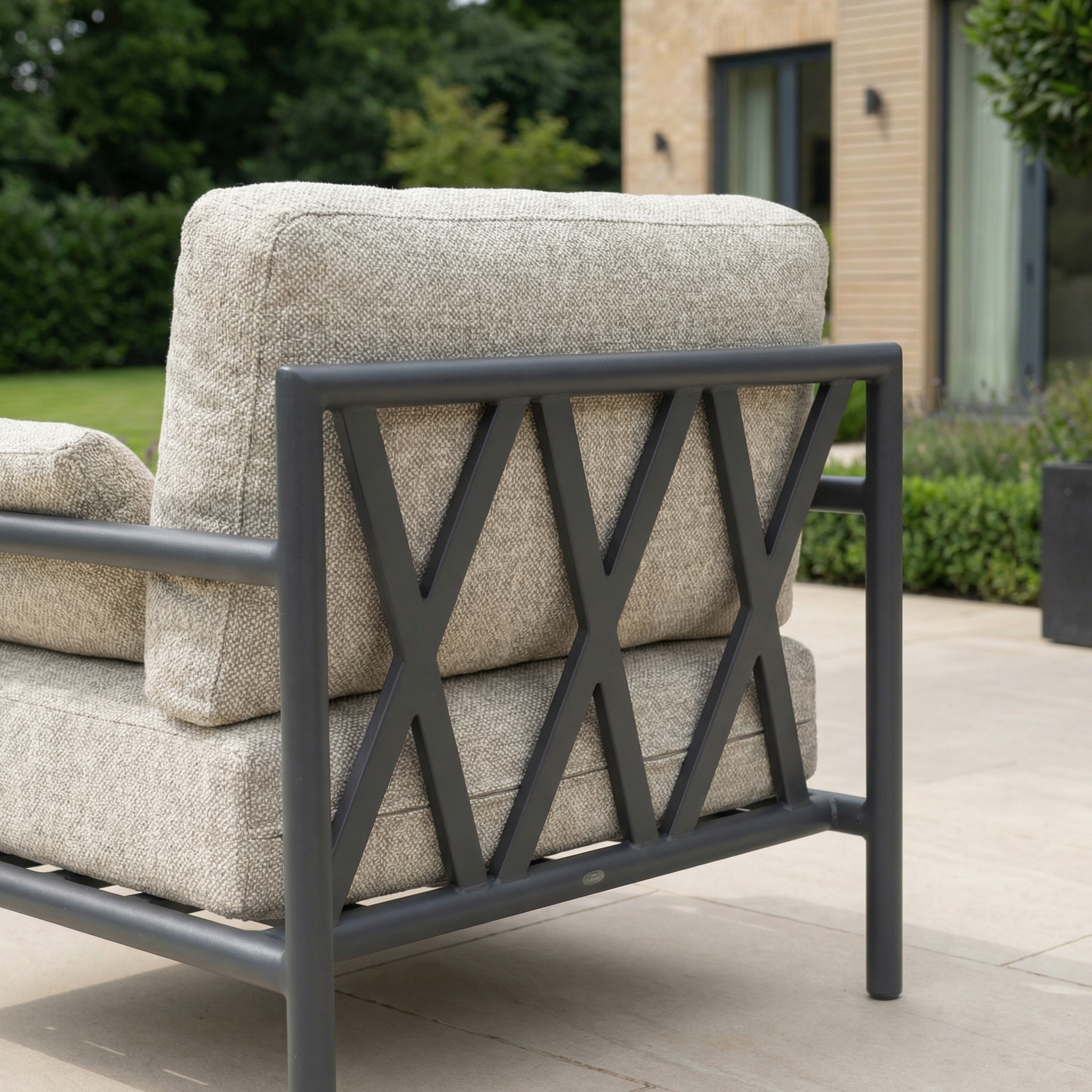 A chair from the Sorelli 2 Seat Sofa Set in Truffle features a gray metal frame and beige cushions, adding stylish comfort to a tiled patio beside a modern building.