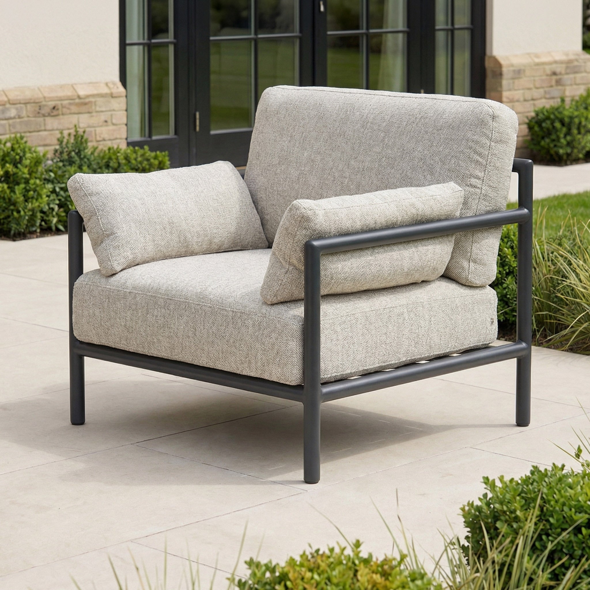 The Sorelli Armchair in Truffle with gray cushions and a black metal frame enhances your patio near greenery, providing a stylish and comfortable outdoor seating option.