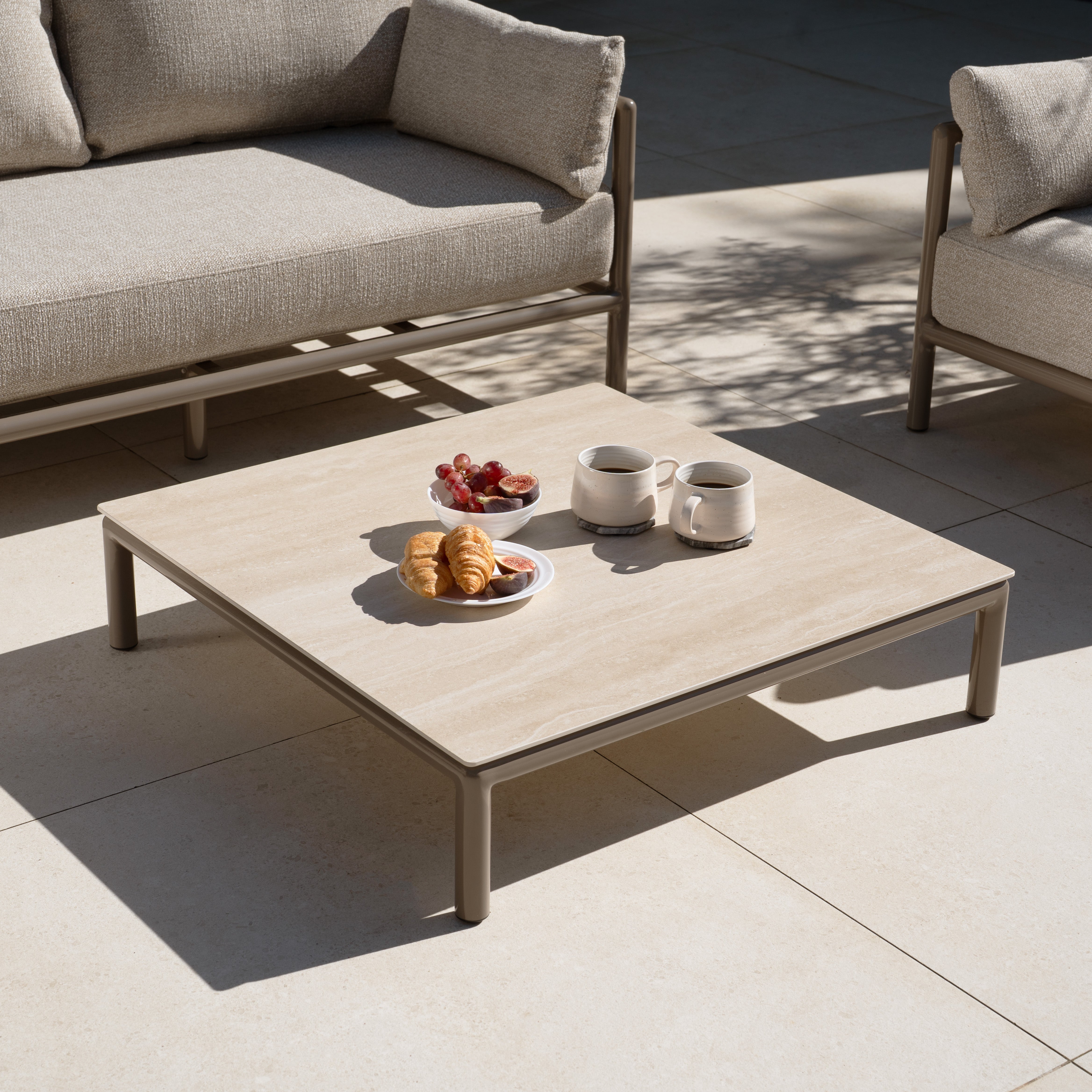 A coffee table with mugs, croissants, and grapes stands between two beige Sorelli 2 Seat Sofa Set in Oatmeal sofas, creating a cozy relaxation spot with weather-resistant garden furniture on the tiled patio.