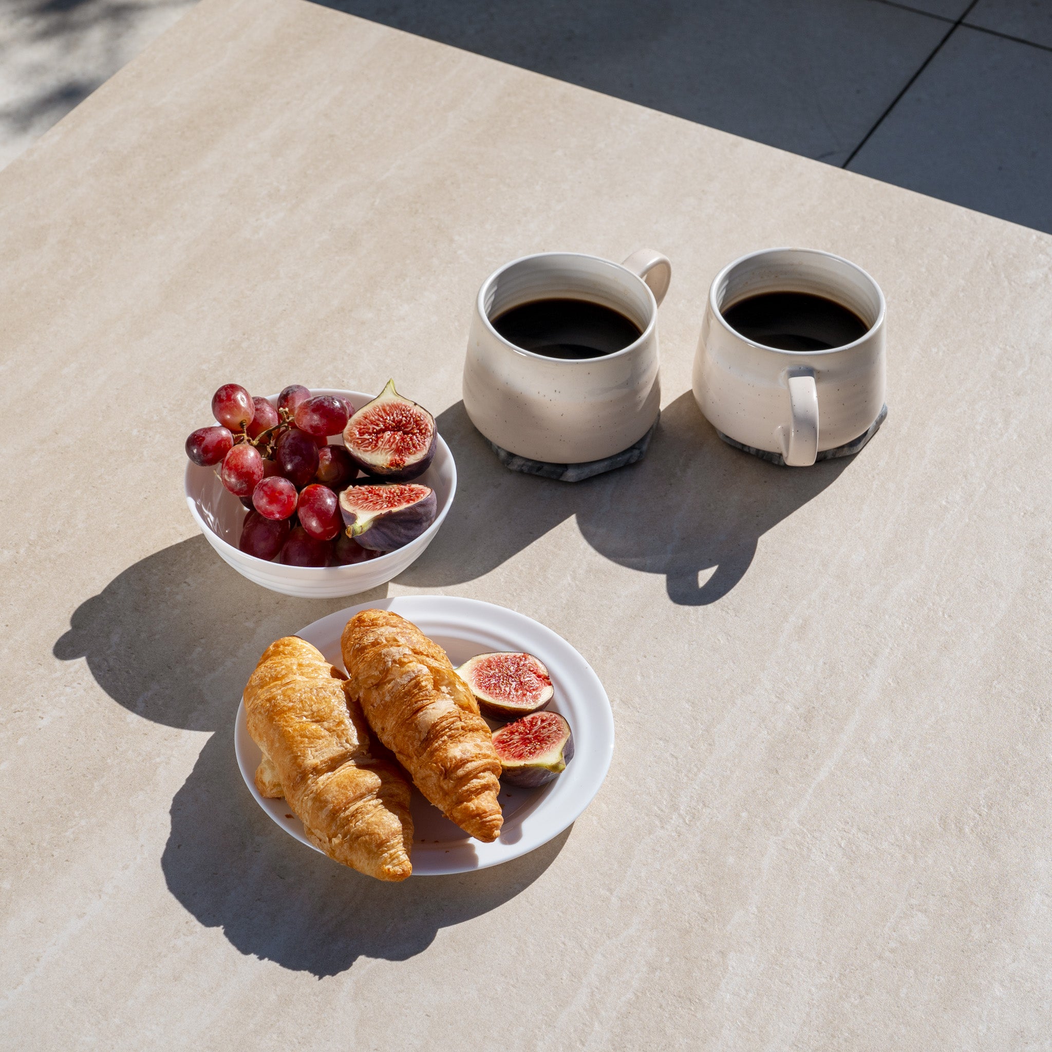 Two cups of black coffee, croissants, figs, and grapes rest on a sunlit table beside the Sorelli 2 Seat Sofa Set in Oatmeal—perfect for enjoying breakfast with stylish, weather-resistant garden furniture.