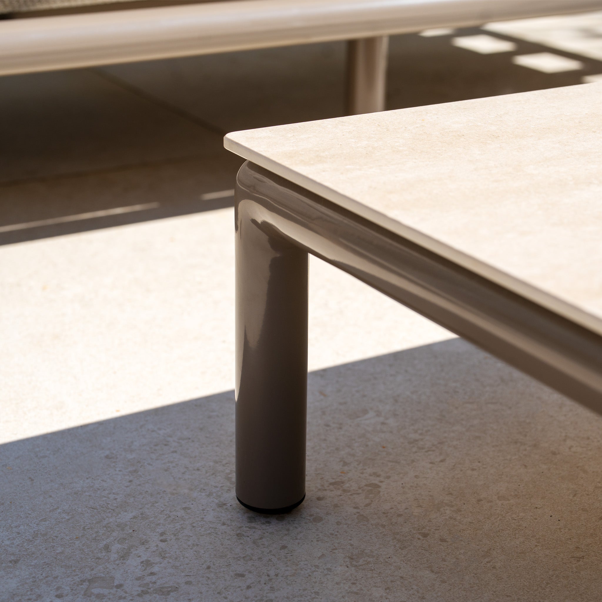 Close-up of a beige metal leg from the Sorelli 2 Seat Sofa Set in Oatmeal, casting a shadow on light concrete in sunlight.