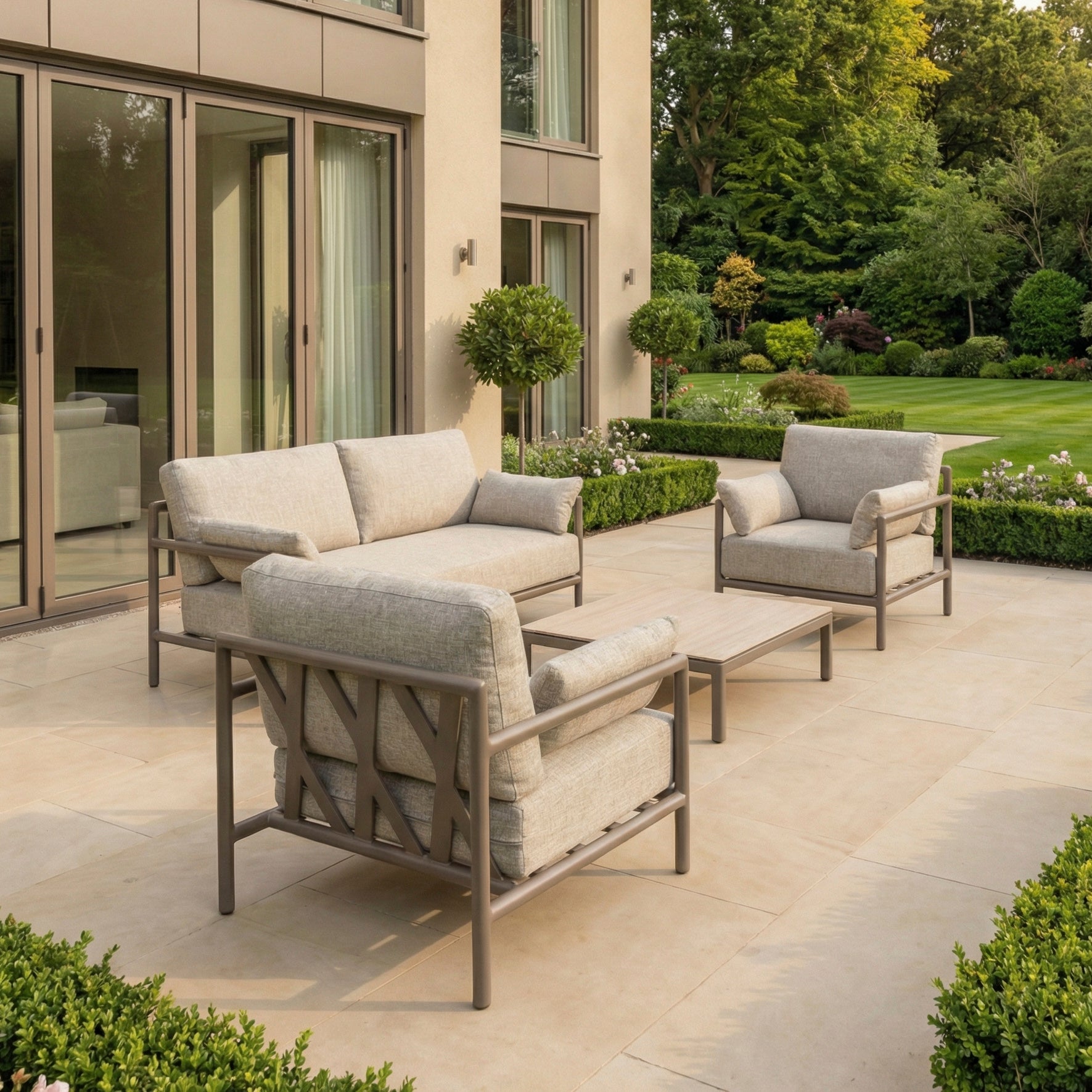 Modern outdoor patio with weather-resistant garden furniture, including a Sorelli 2 Seat Sofa Set in Oatmeal with cushioned seating, a coffee table, and a landscaped garden in the background.