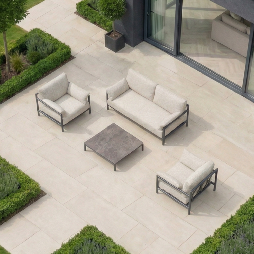 Modern outdoor patio with the Sorelli 2 Seat Sofa Set in Truffle, including two chairs and a low table on a tiled terrace by a lush garden—perfect for relaxing in style.