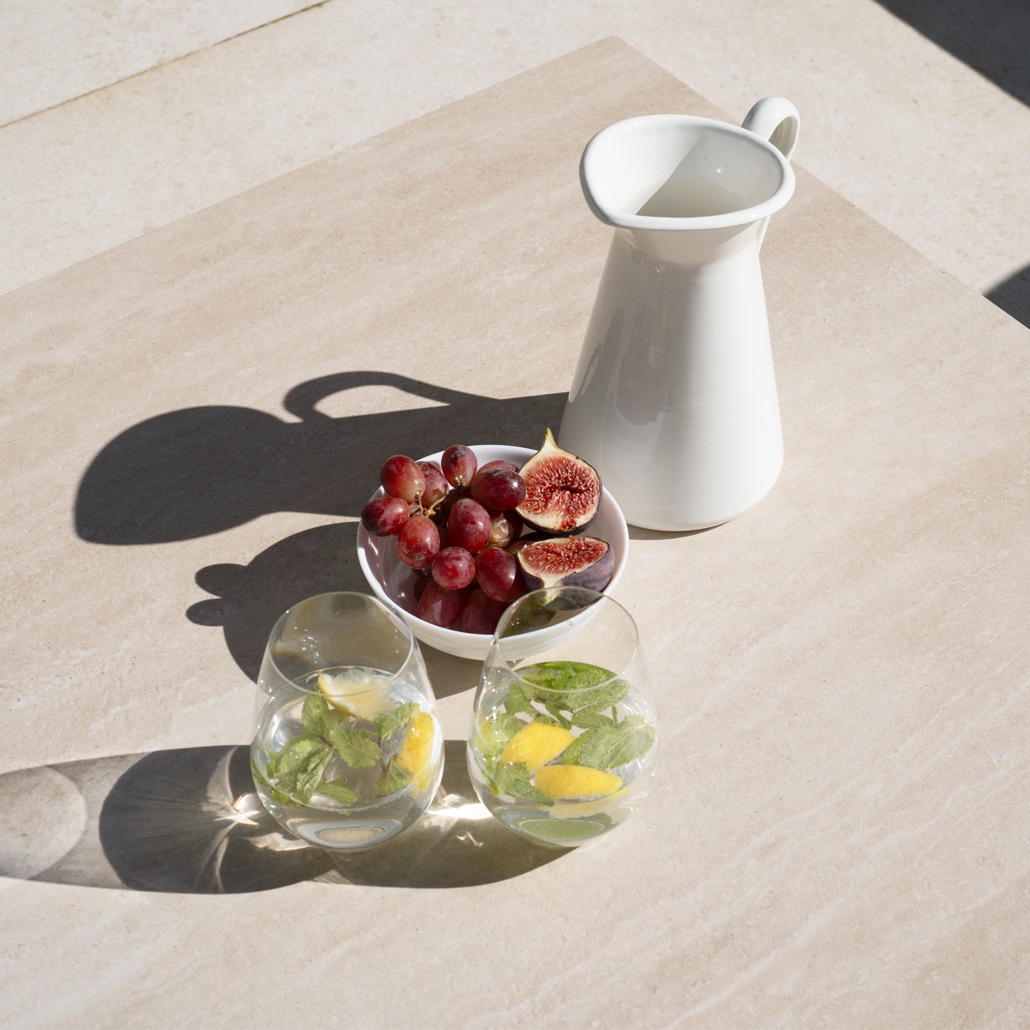 A white pitcher, a bowl of grapes and figs, and two glasses of water with lemon and mint rest elegantly on a beige table paired with the Sorelli 3 Seat Sofa Set in Oatmeal.