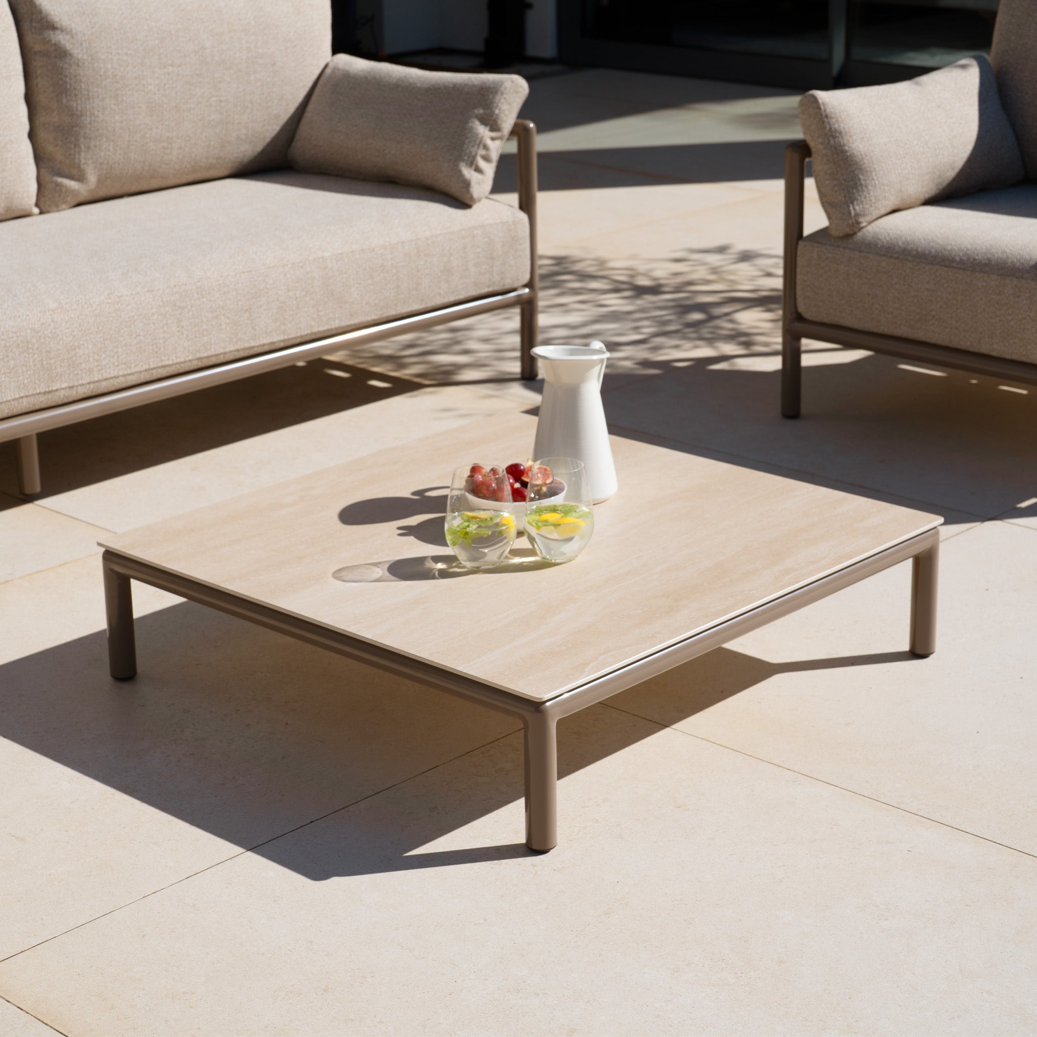 A low coffee table with drinks, berries, and a pitcher stands between two beige Sorelli 3 Seat Sofas in Oatmeal, creating a cozy patio retreat.