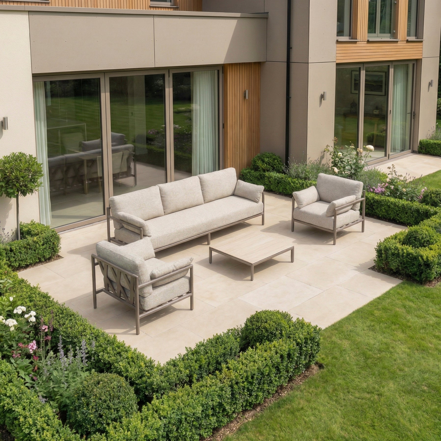 Modern outdoor patio featuring the Sorelli 3 Seat Sofa Set in Oatmeal—a weather-resistant set with a beige sofa, two armchairs, and a table, all surrounded by manicured green bushes.