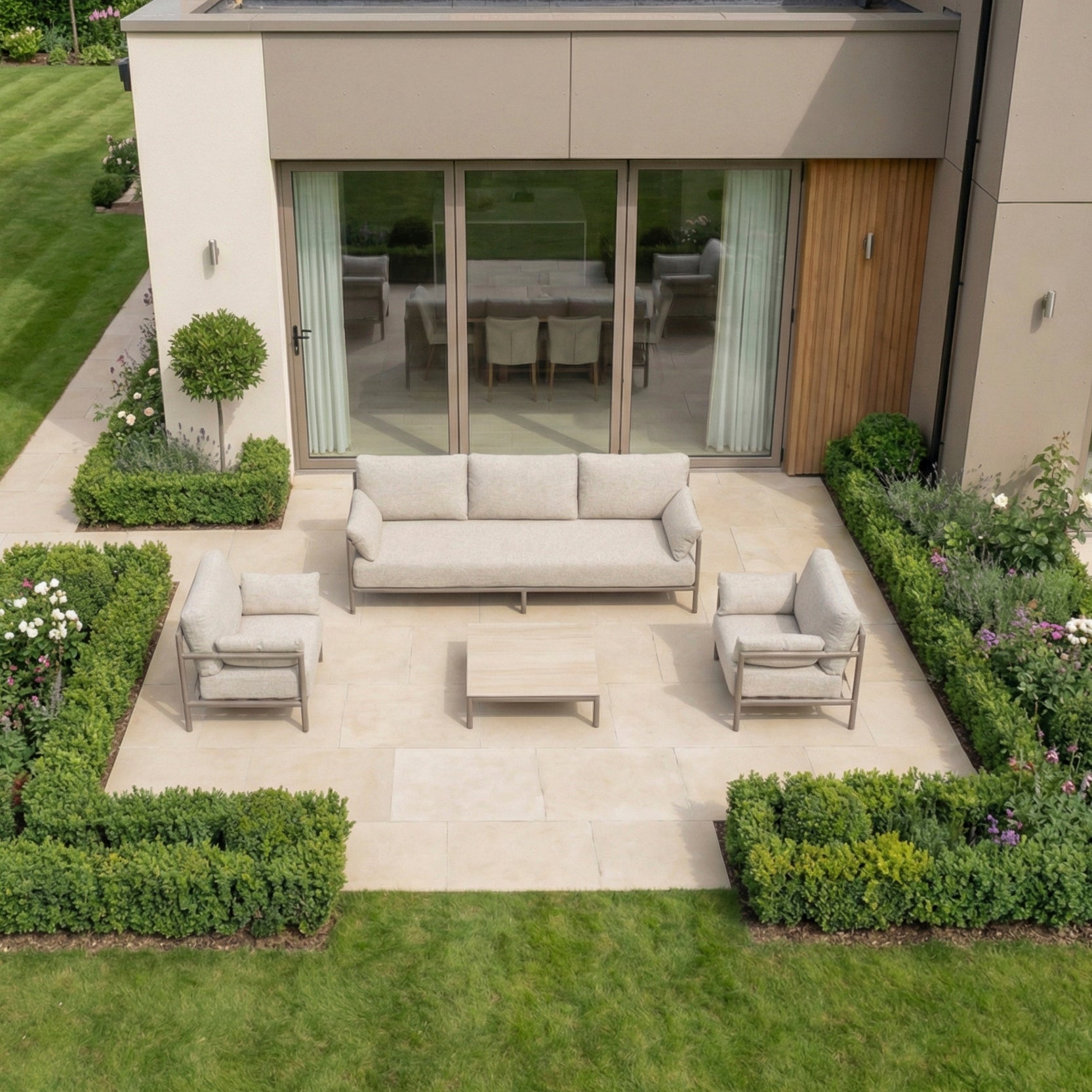 Modern outdoor patio with the Sorelli 3 Seat Sofa Set in Oatmeal, surrounded by green hedges and plants next to a house with glass doors.