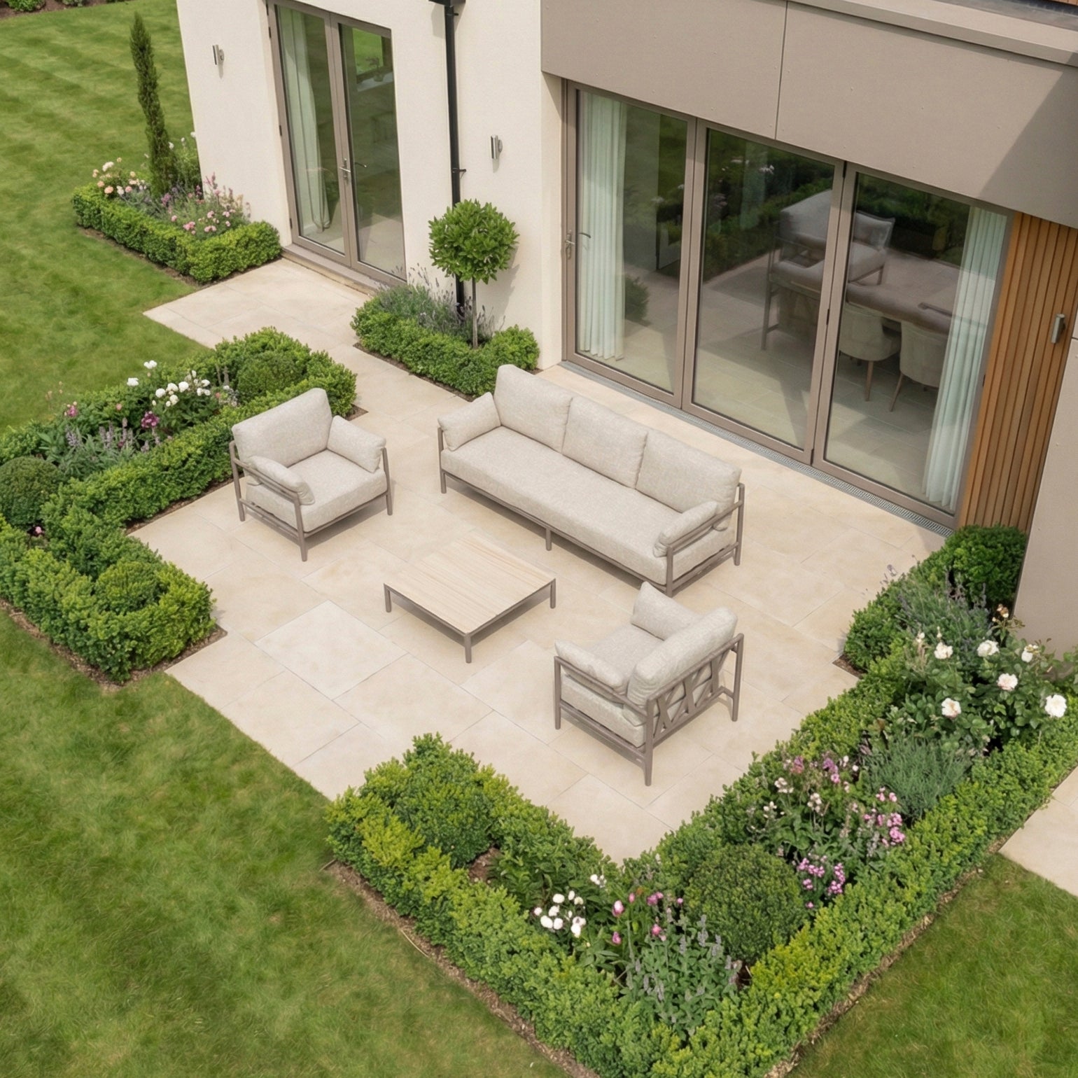 Modern outdoor patio with the Sorelli 3 Seat Sofa Set in Oatmeal, a weather-resistant set featuring beige sofas, a coffee table, and lush green shrubs and flowers all around.