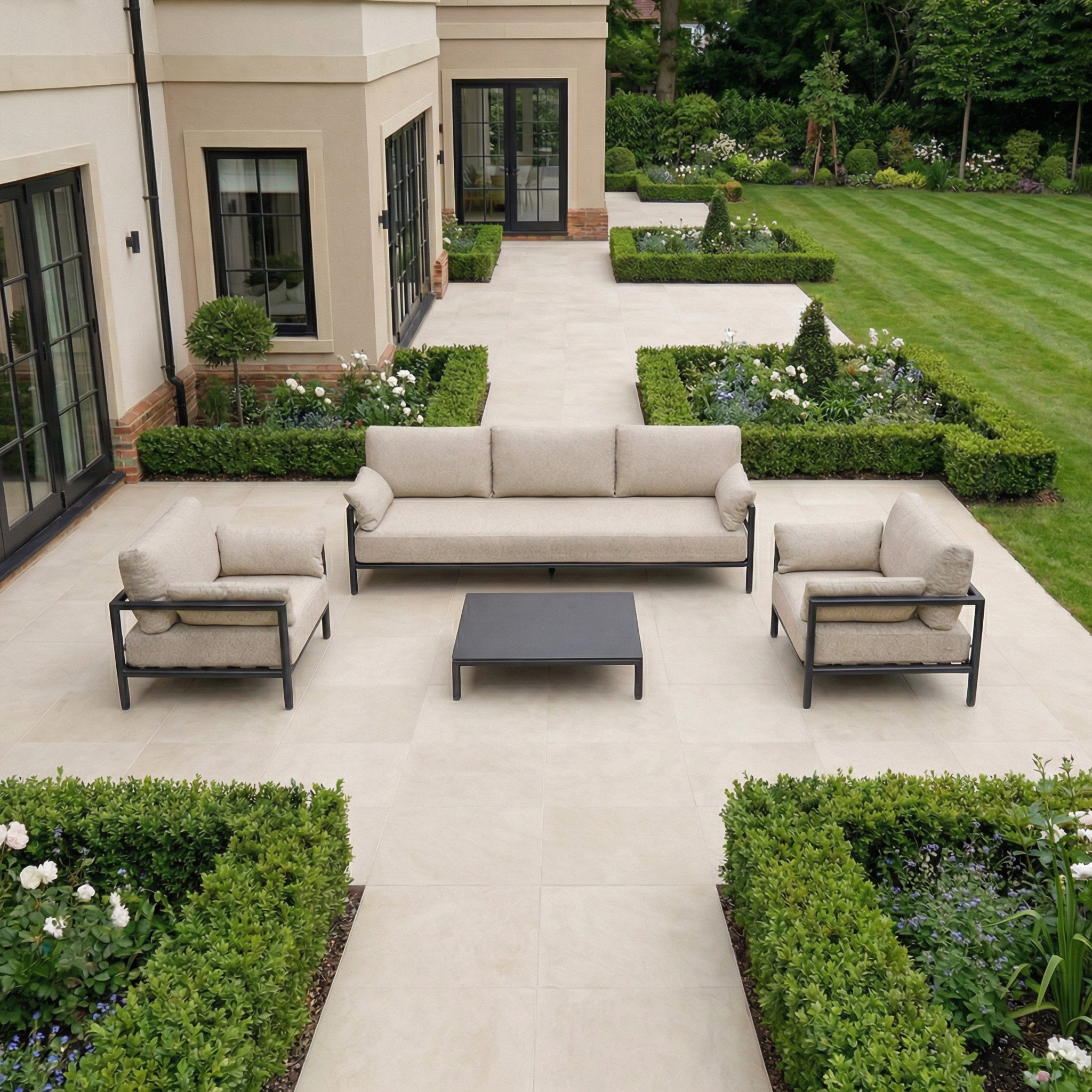 The Sorelli 3 Seat Sofa Set in Truffle offers stylish outdoor seating with chairs and table, ideal for relaxing on a tiled terrace surrounded by a beautifully manicured garden.