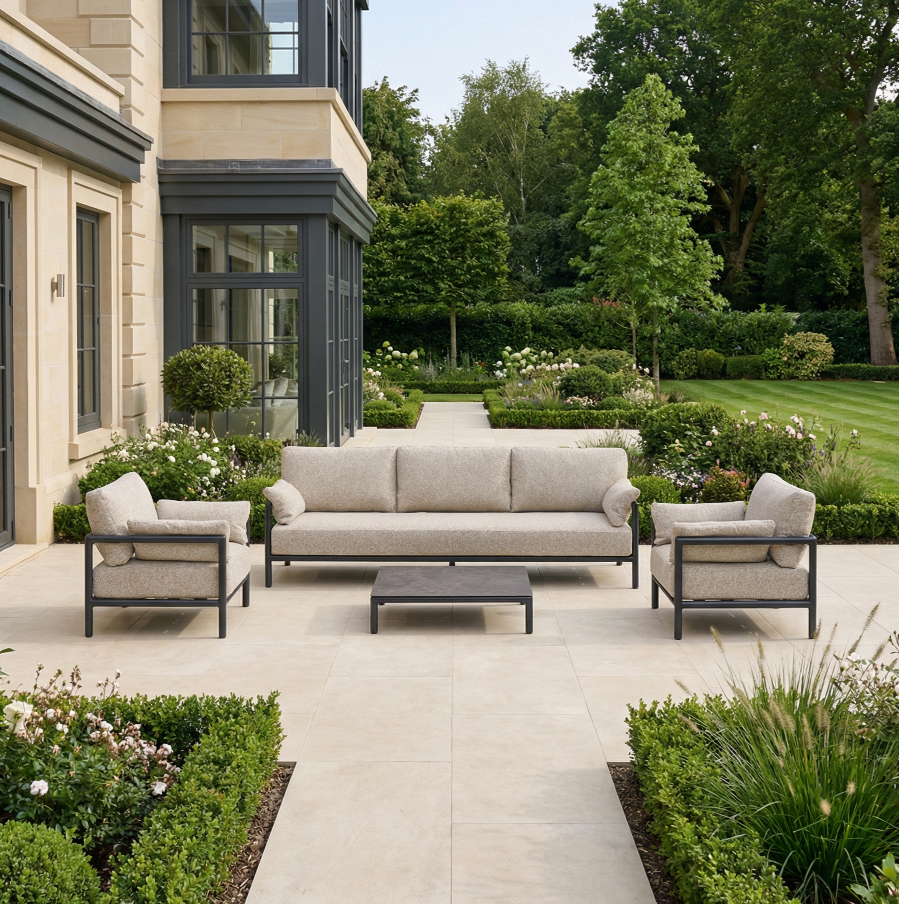 Outdoor patio featuring the Sorelli 3 Seat Sofa Set in Truffle, matching armchairs, and a low table—ideal garden furniture for chic and comfortable outdoor seating amid manicured gardens.