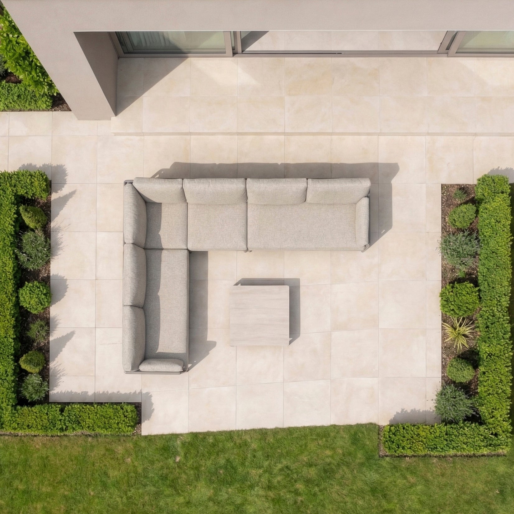 Aerial view of a patio showcasing the Sorelli Corner Sofa Set in Oatmeal, a chic outdoor sofa with aluminium frame and coffee table, surrounded by lush green hedges.