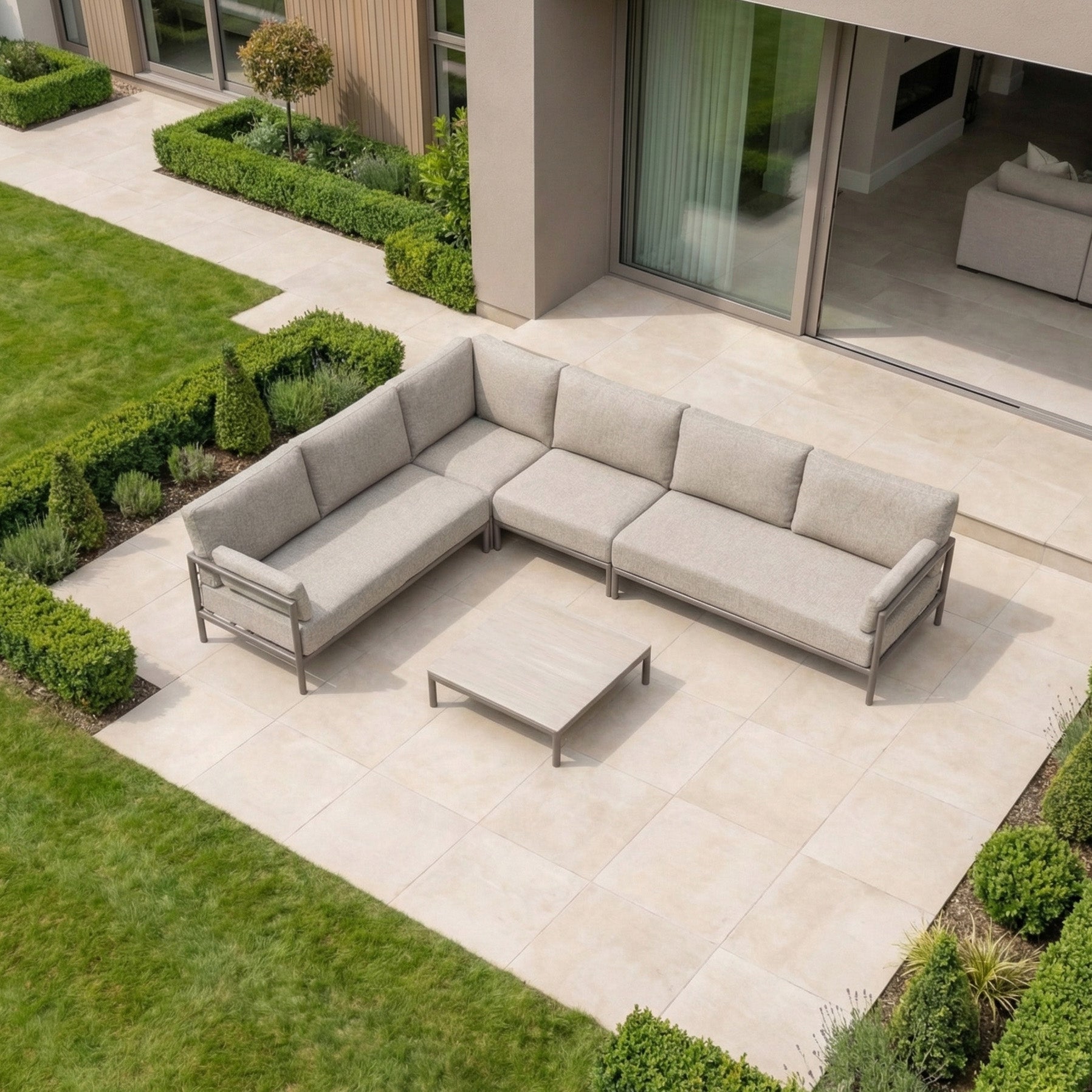 Outdoor patio featuring the Sorelli Corner Sofa Set in Oatmeal with a sturdy aluminium frame, beige cushions, and coffee table on tiled flooring, surrounded by green grass and shrubs.