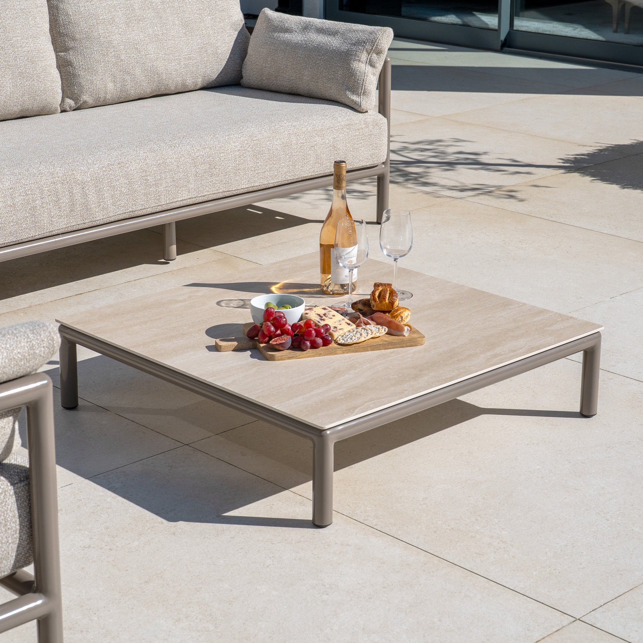 A low patio table with wine, glasses, fruit, and snacks sits beside the Sorelli Corner Sofa Set in Oatmeal, featuring a sleek aluminium frame on a tiled terrace.