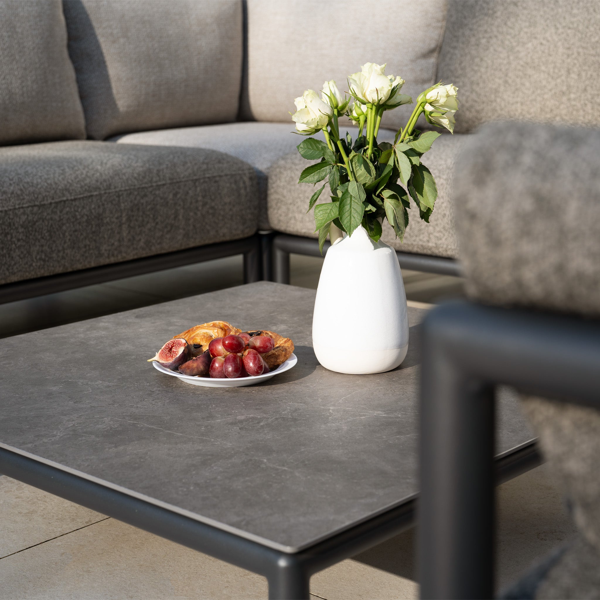 A white vase with white roses and a plate of fruit sit on a gray coffee table, with the Sorelli Corner Sofa Set in Truffle adding modern charm to your garden furniture in the background.