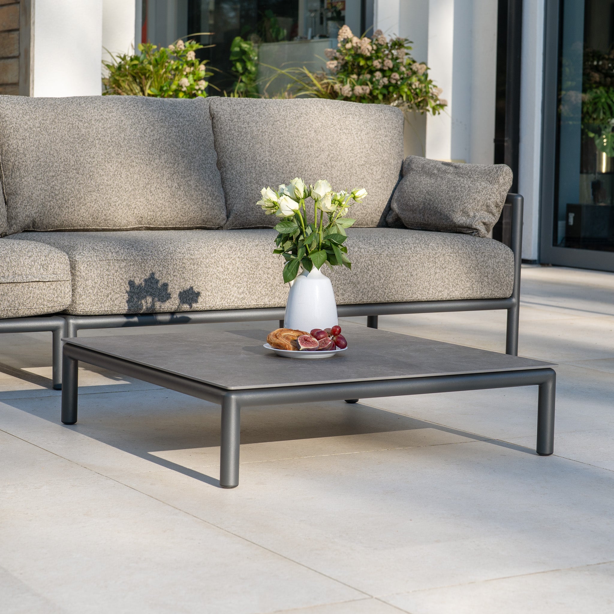 The Sorelli Corner Sofa Set in Truffle and a low table with a vase of white flowers, pastries, and grapes create the perfect garden furniture for relaxing on a sunny patio.