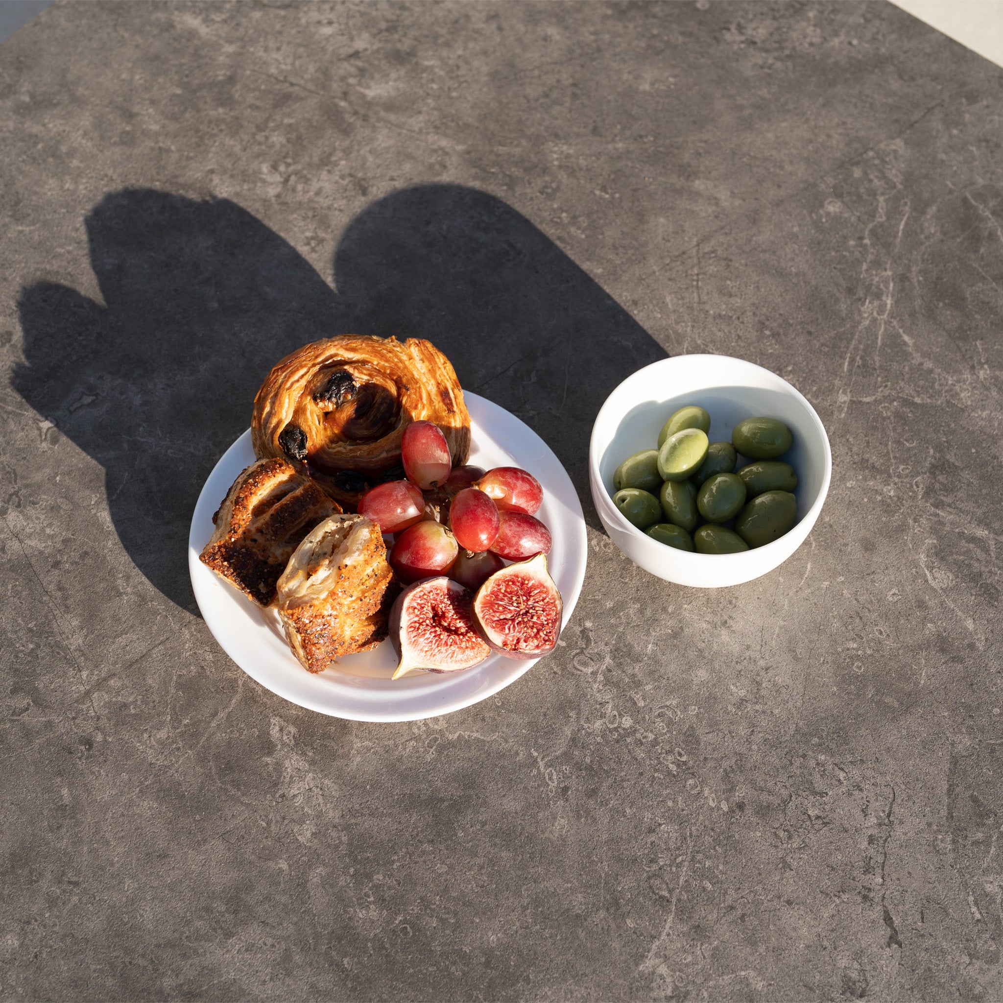 A plate with pastries, figs, and grapes sits beside a bowl of green olives on a gray table, perfect for enjoying atop your Sorelli U Shape Corner Sofa Set in Truffle.