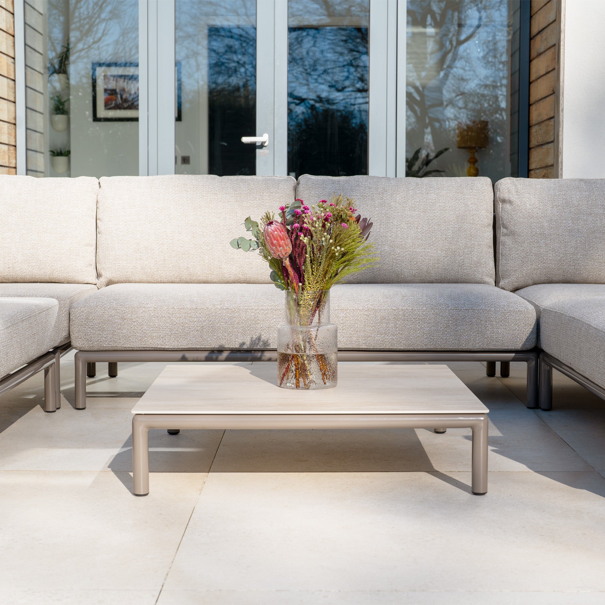 A glass vase with flowers sits on a low table between beige outdoor sofas from the Sorelli U Shape Corner Sofa Set in Oatmeal on a patio.
