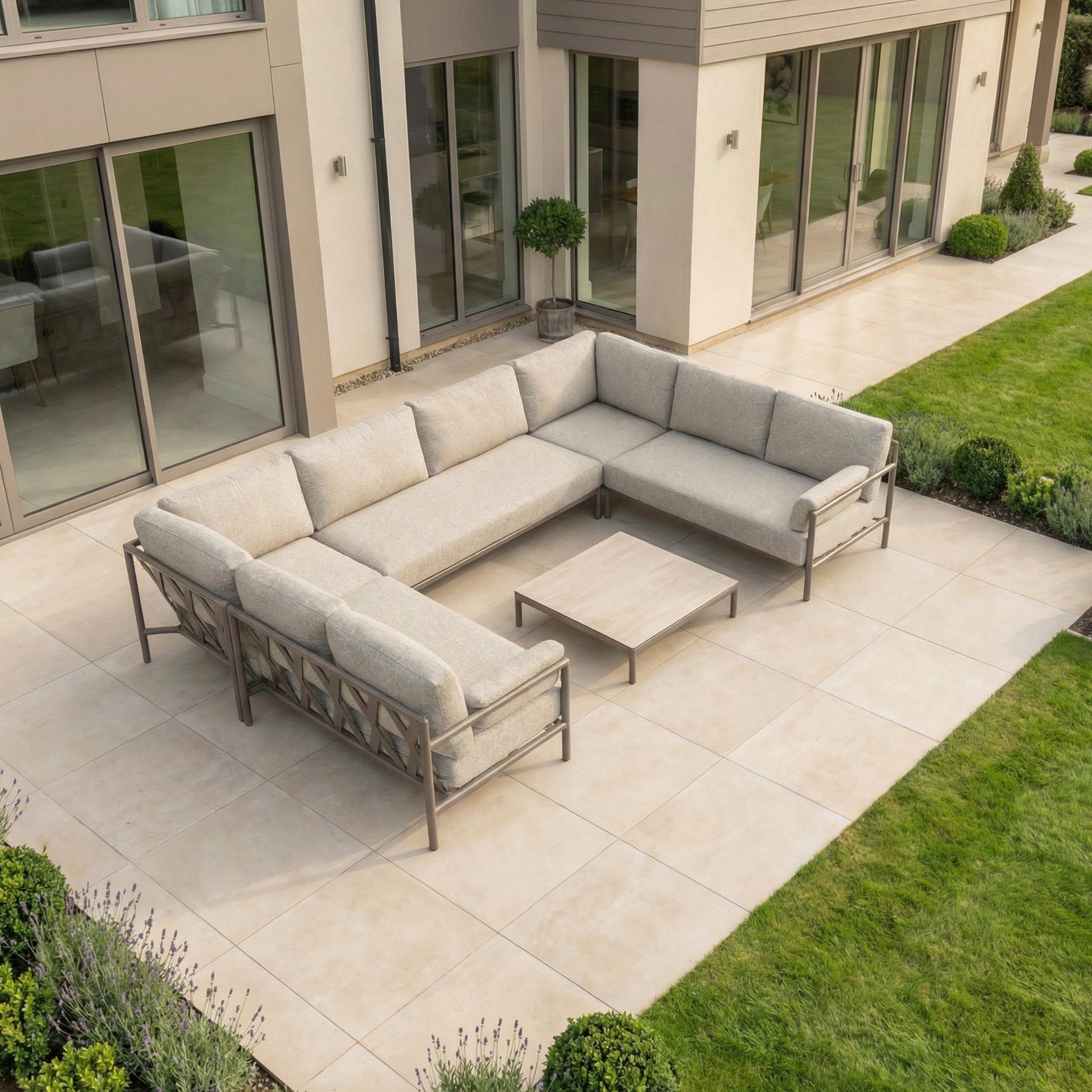 Outdoor patio with the Sorelli U Shape Corner Sofa Set in Oatmeal, featuring an aluminum frame and low table on light tiles, next to a modern house and green lawn.